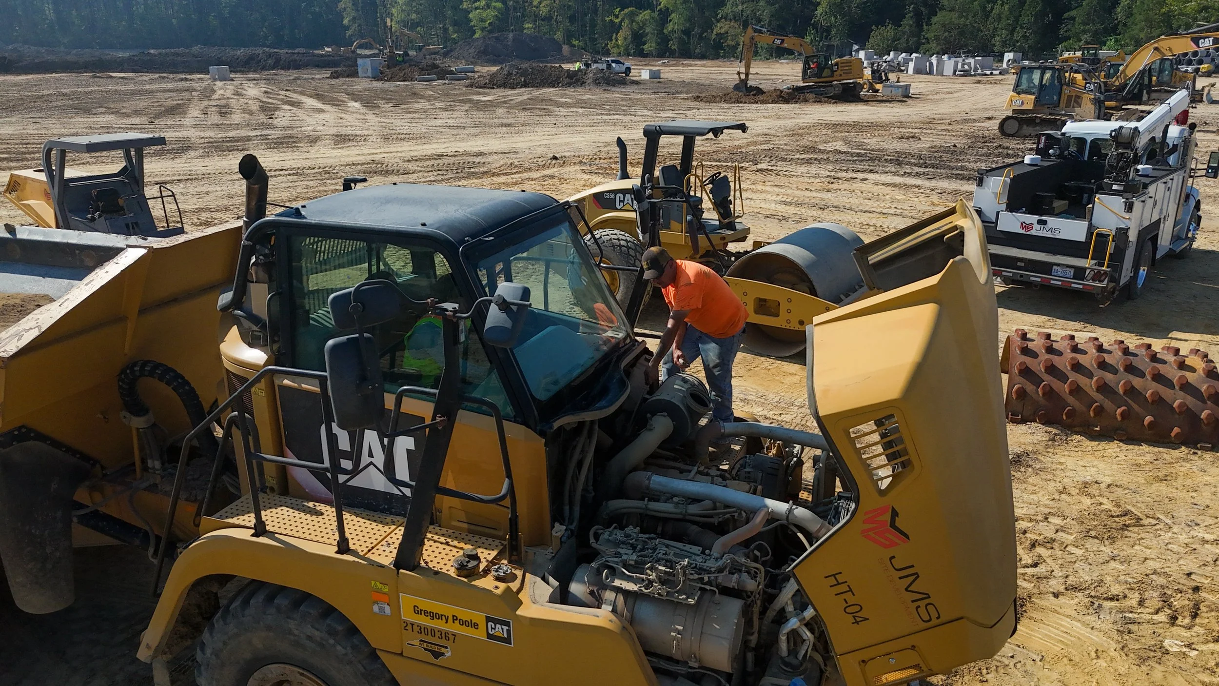 Construction site with multiple heavy machinery including bulldozers and excavators, with a worker in orange shirt working near a yellow Caterpillar vehicle.
