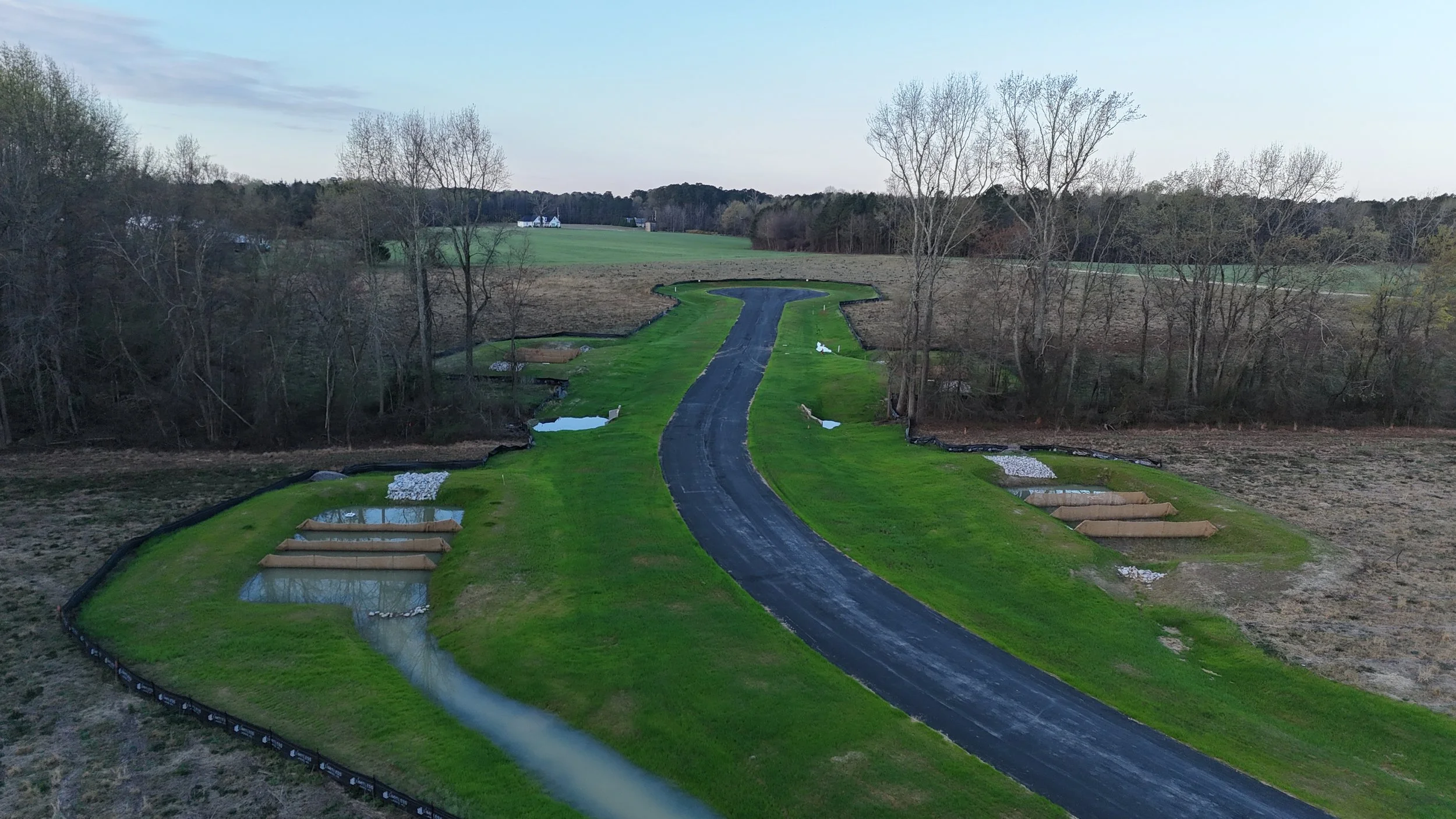 A golf course under construction with a paved cart path winding through green grass, dirt patches, and several small water hazards, surrounded by trees and open fields in the background.