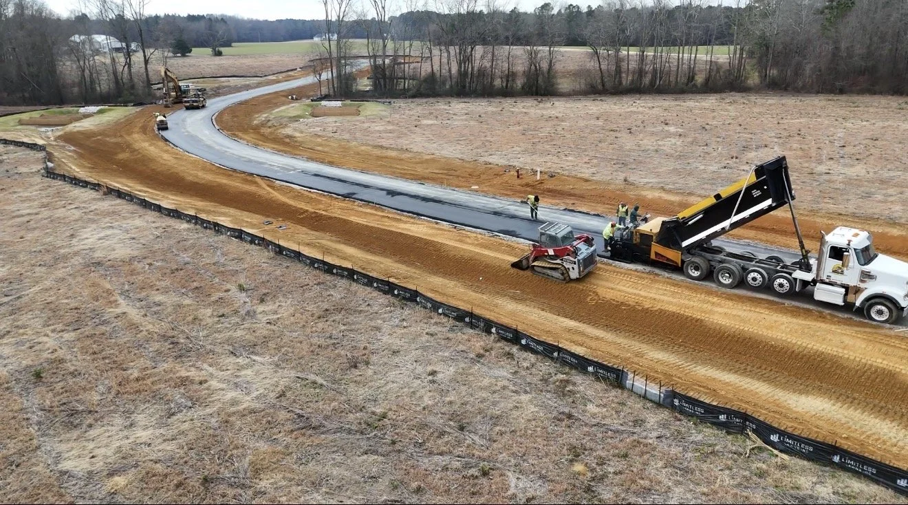Workers and construction vehicles paving a new winding road through a rural area with fields and trees.
