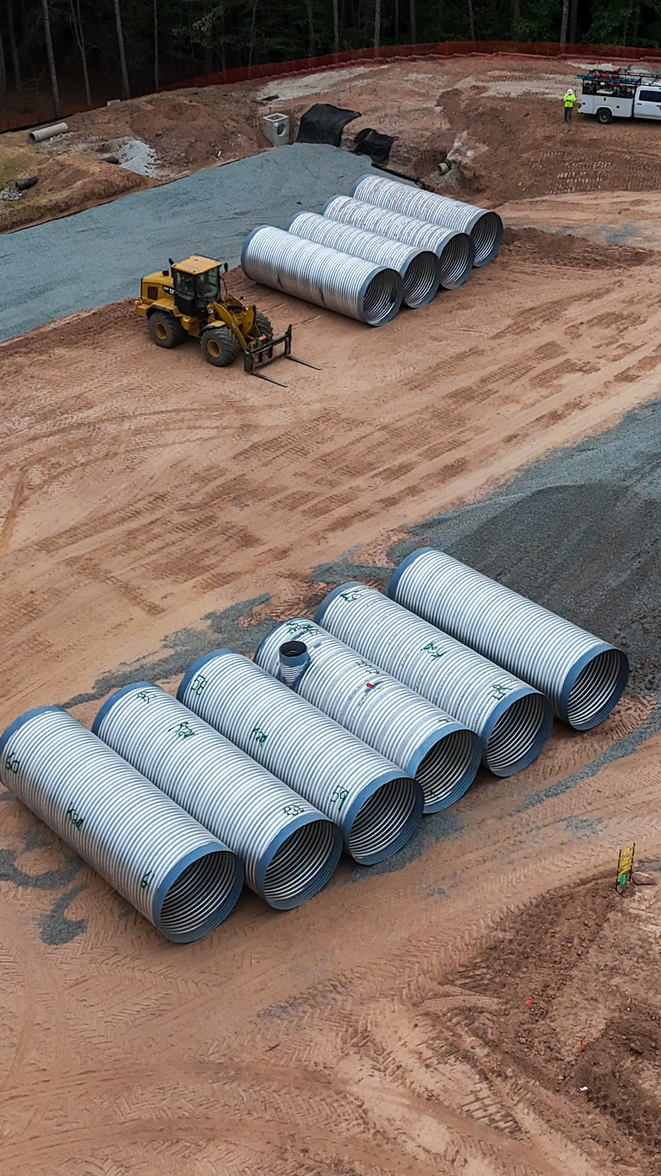 Construction site with large metal pipes, a bulldozer, and a worker in a yellow vest near a white utility truck. The area is dug out with dirt and gravel.