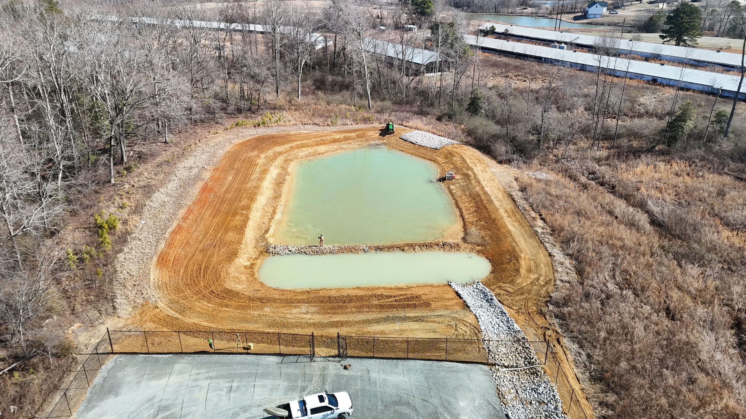 Aerial view of a construction site with two small ponds, one with light green water and the other with pale blue water. The site is surrounded by trees and some construction equipment, including a small roller and several workers. A gravel path runs alongside the ponds, and a fence borders part of the site with a parked white truck in the lower part of the image.