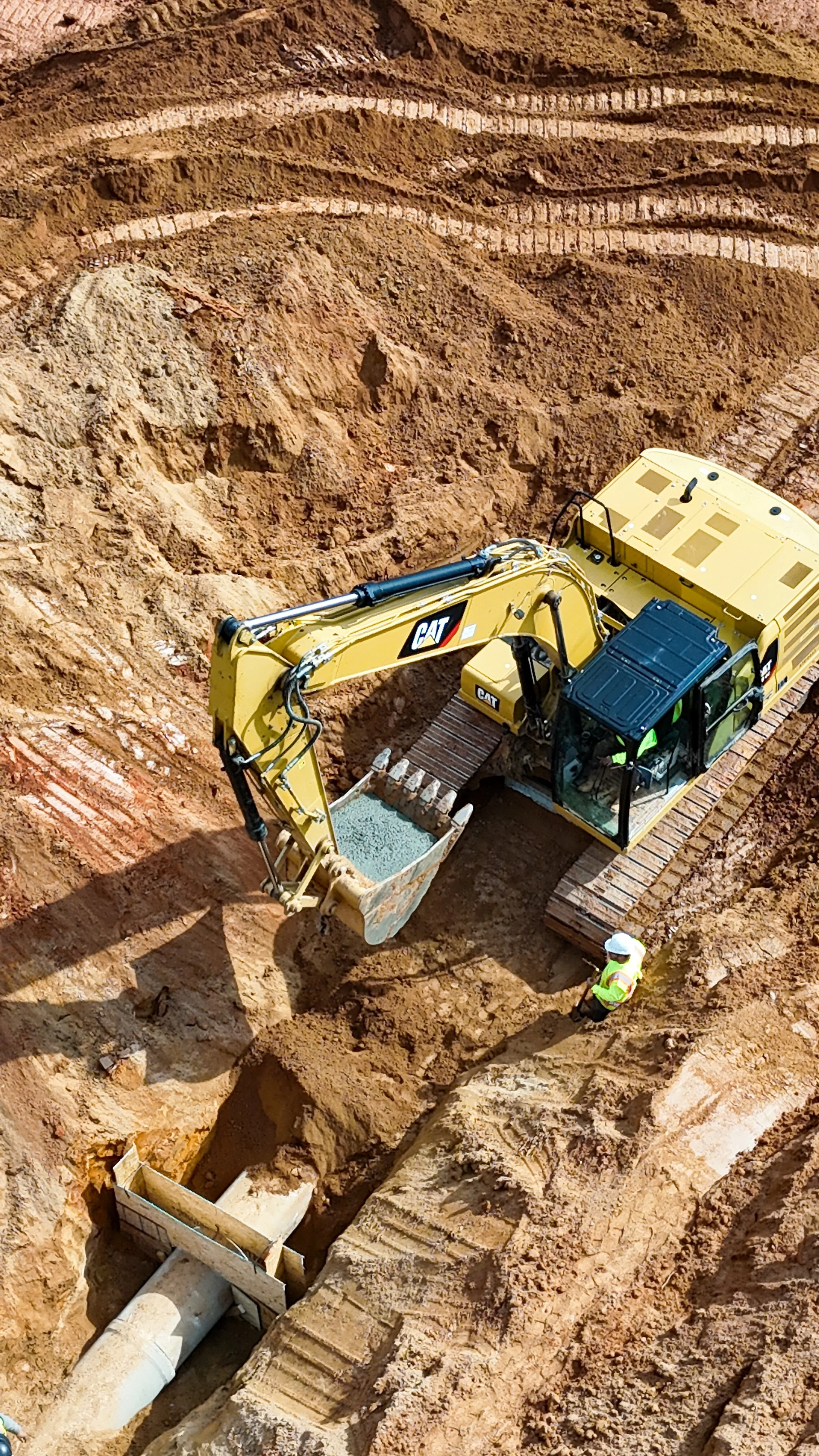 An excavator is digging a trench in a construction site with two workers overseeing the work, and a large concrete pipe being installed.