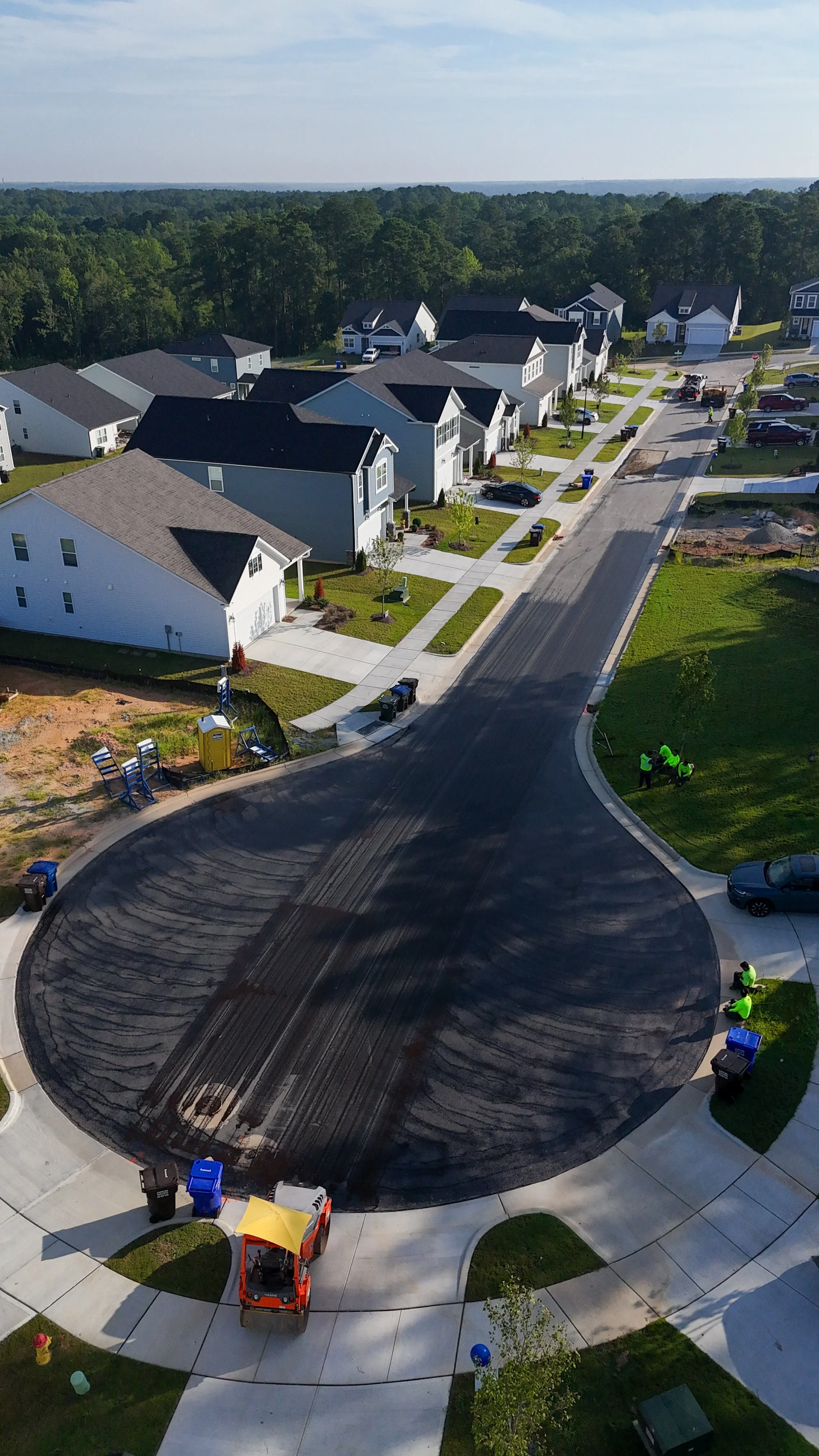 An aerial view of a neighborhood with houses, a newly paved road, construction workers, and equipment installing asphalt.