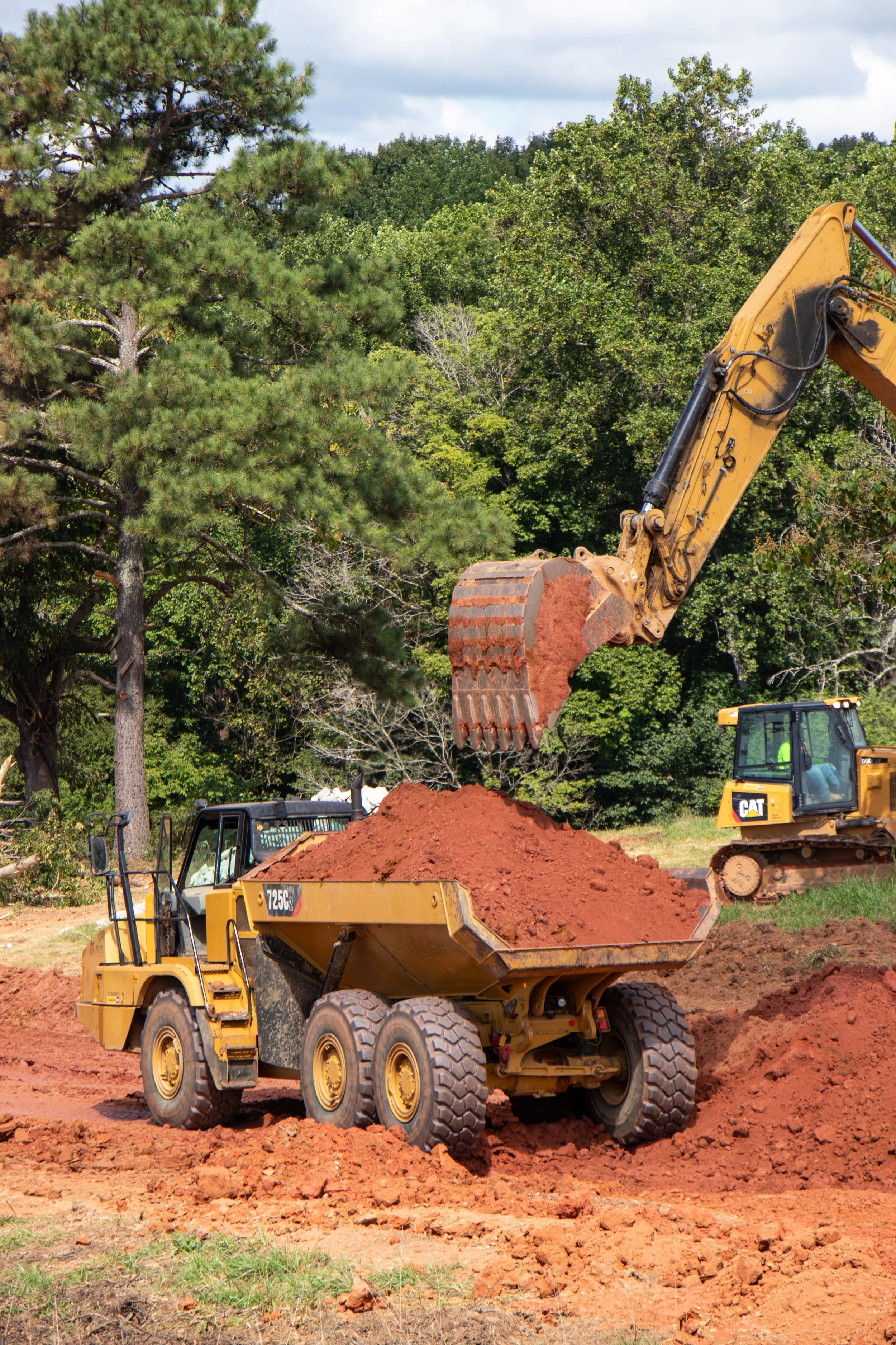 Construction site with a yellow backhoe loader dumping reddish dirt into a dump truck, surrounded by green trees and a cloudy sky.