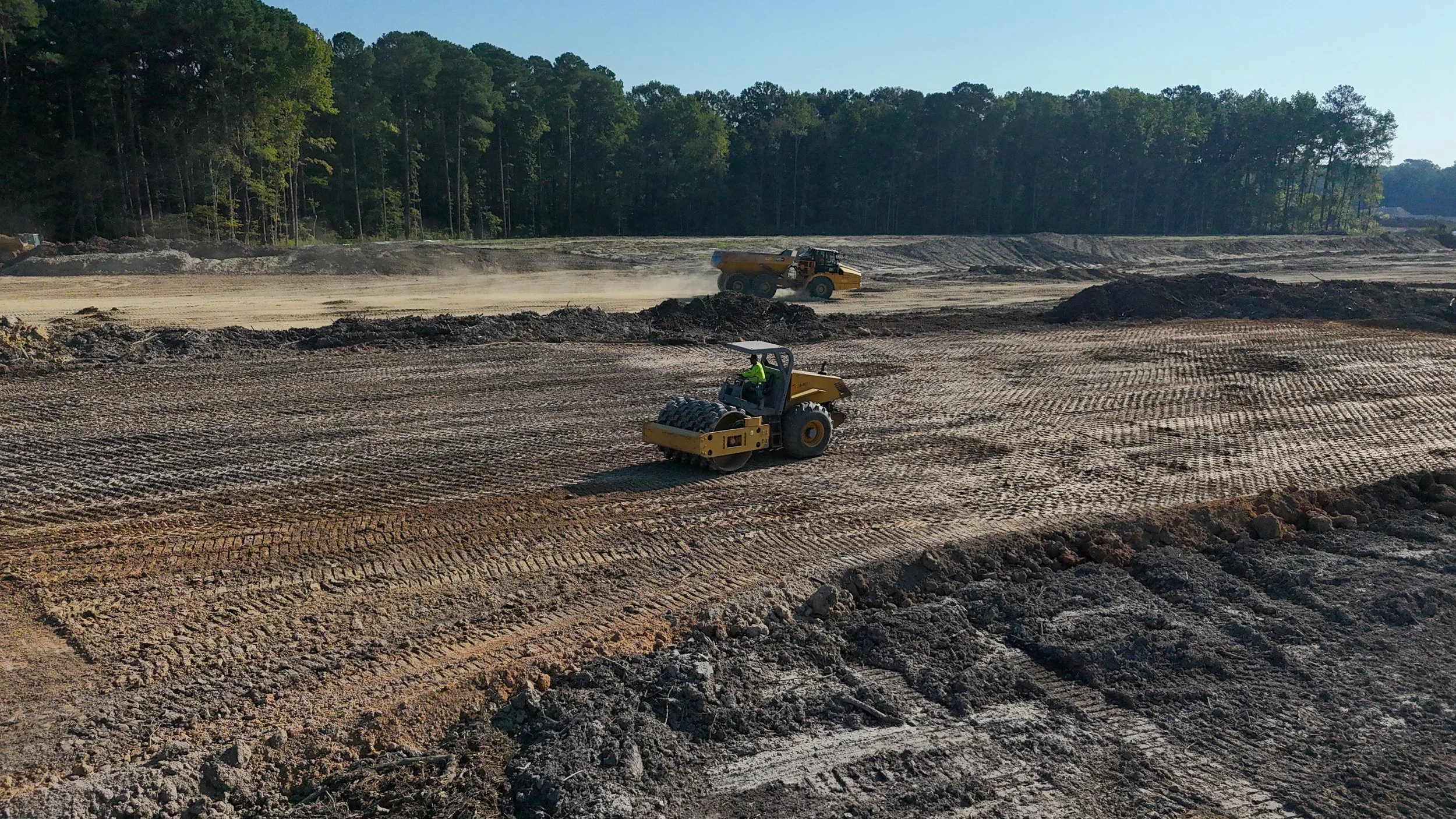 Construction site with two road rollers compacting dirt. Foreground shows freshly graded earth with tire marks. Background features trees and a clear sky.
