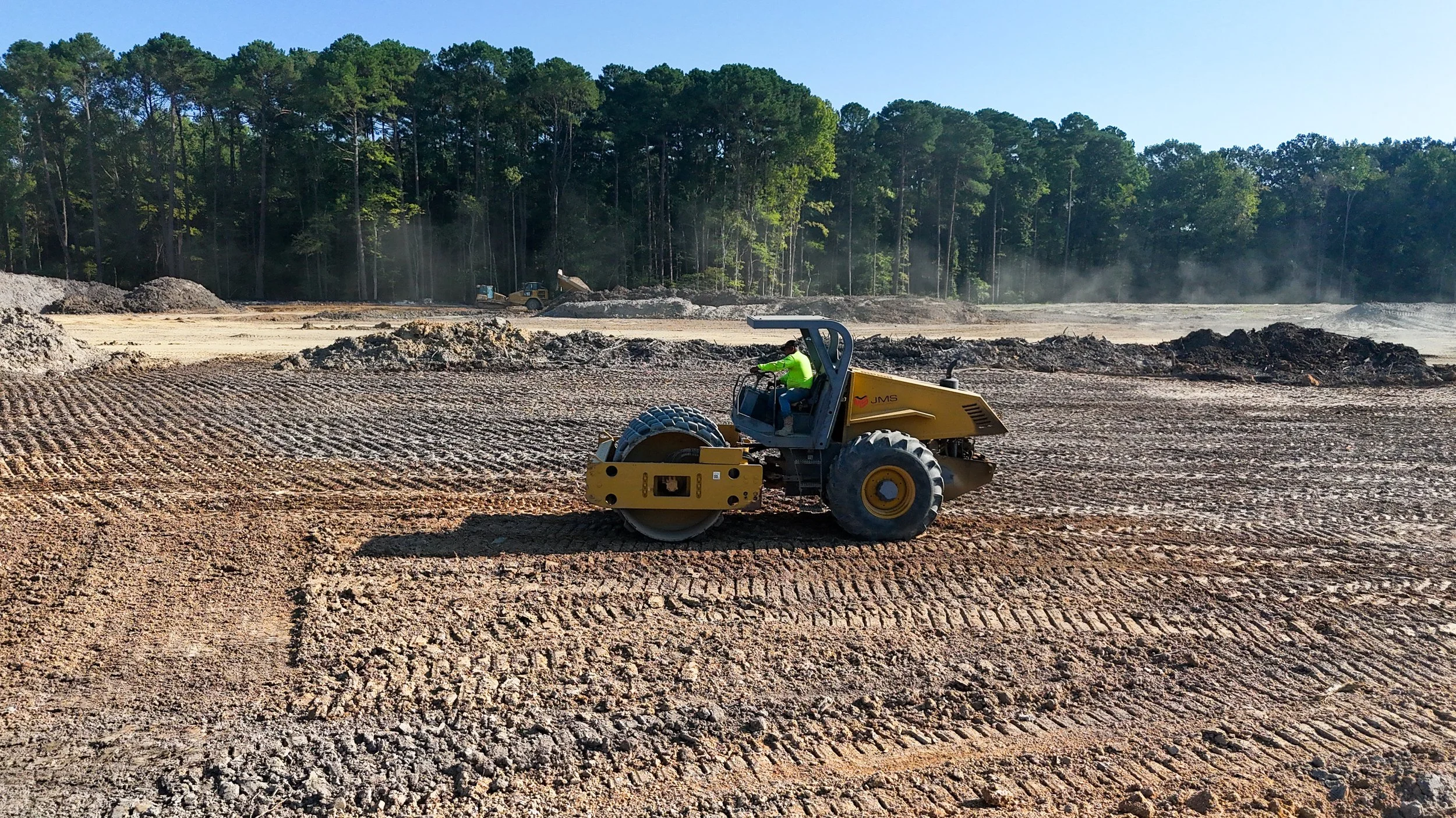 Construction site with a worker operating a yellow road roller, leveling the ground with trees in the background under a clear sky.