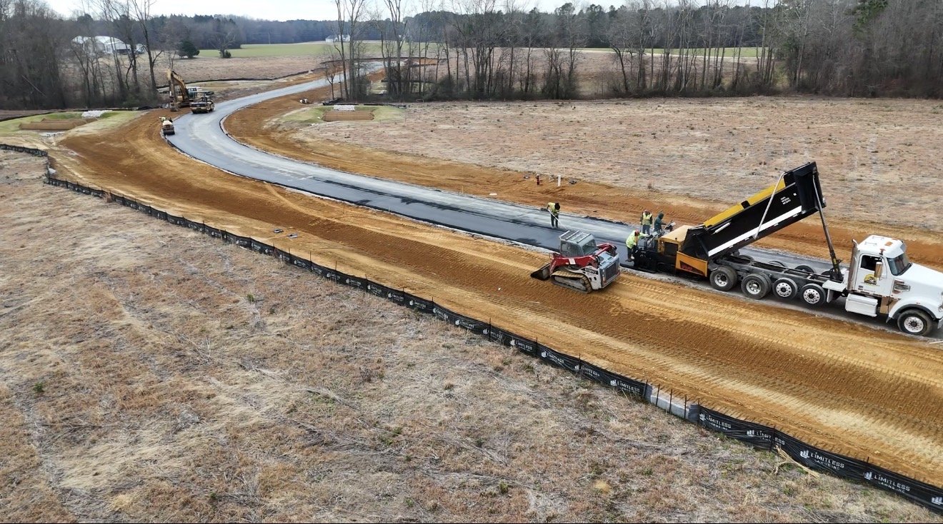 Construction workers paving a curved road in a rural area with dirt and grass surrounding it. Heavy machinery, including a roller and a dump truck, are present on the site.