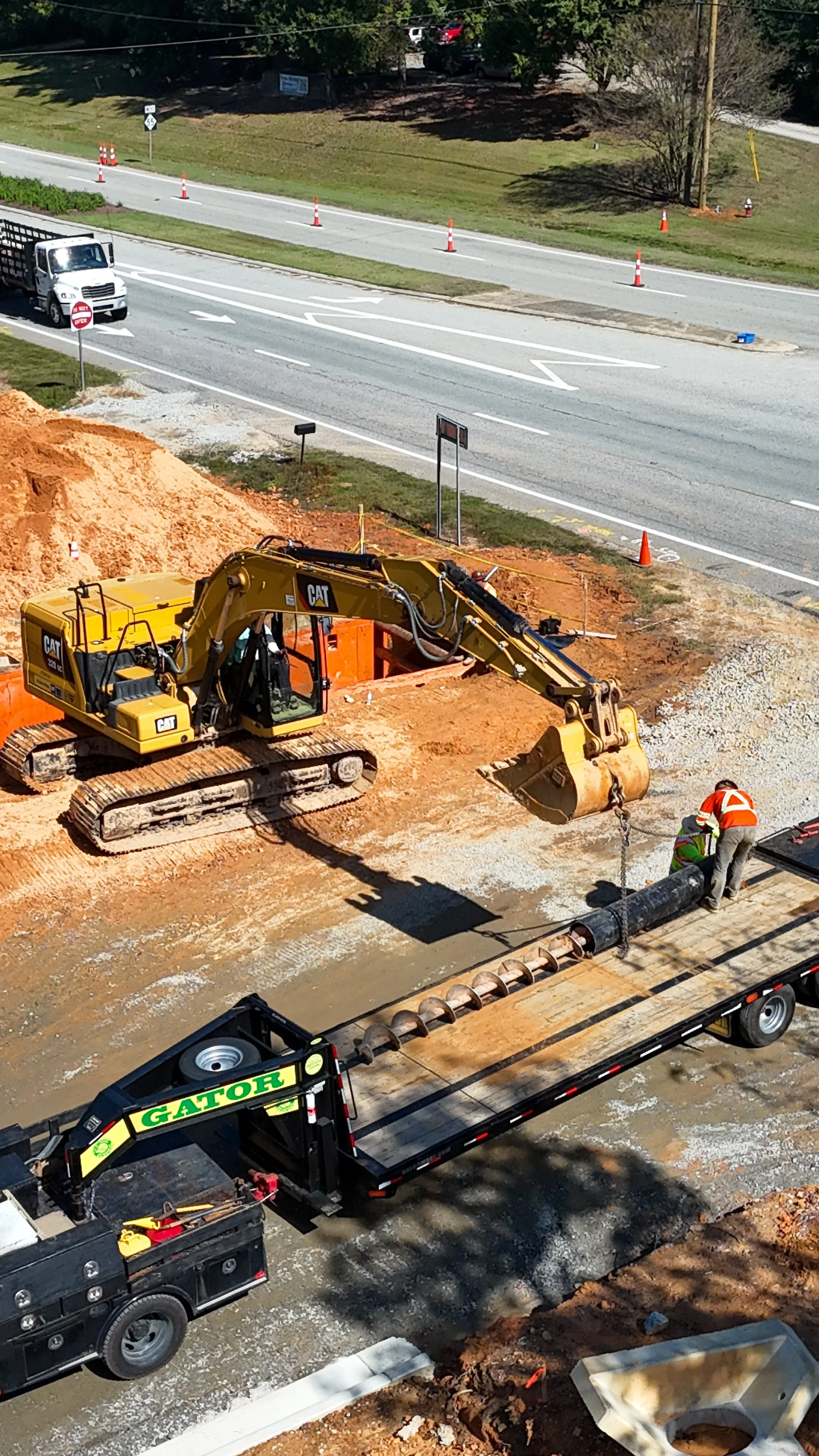Construction workers and heavy machinery work on a road expansion project, with a large CAT excavator digging and moving earth in a partially excavated area, while traffic cones and barriers manage traffic on the nearby road.