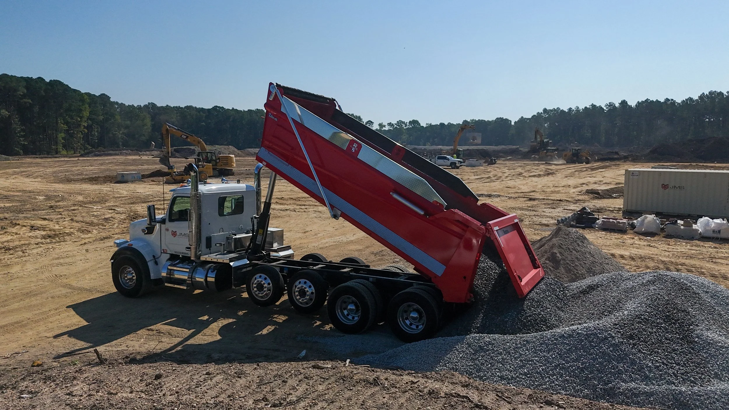 Construction site with a dump truck dumping gravel, with excavators and construction equipment in the background.