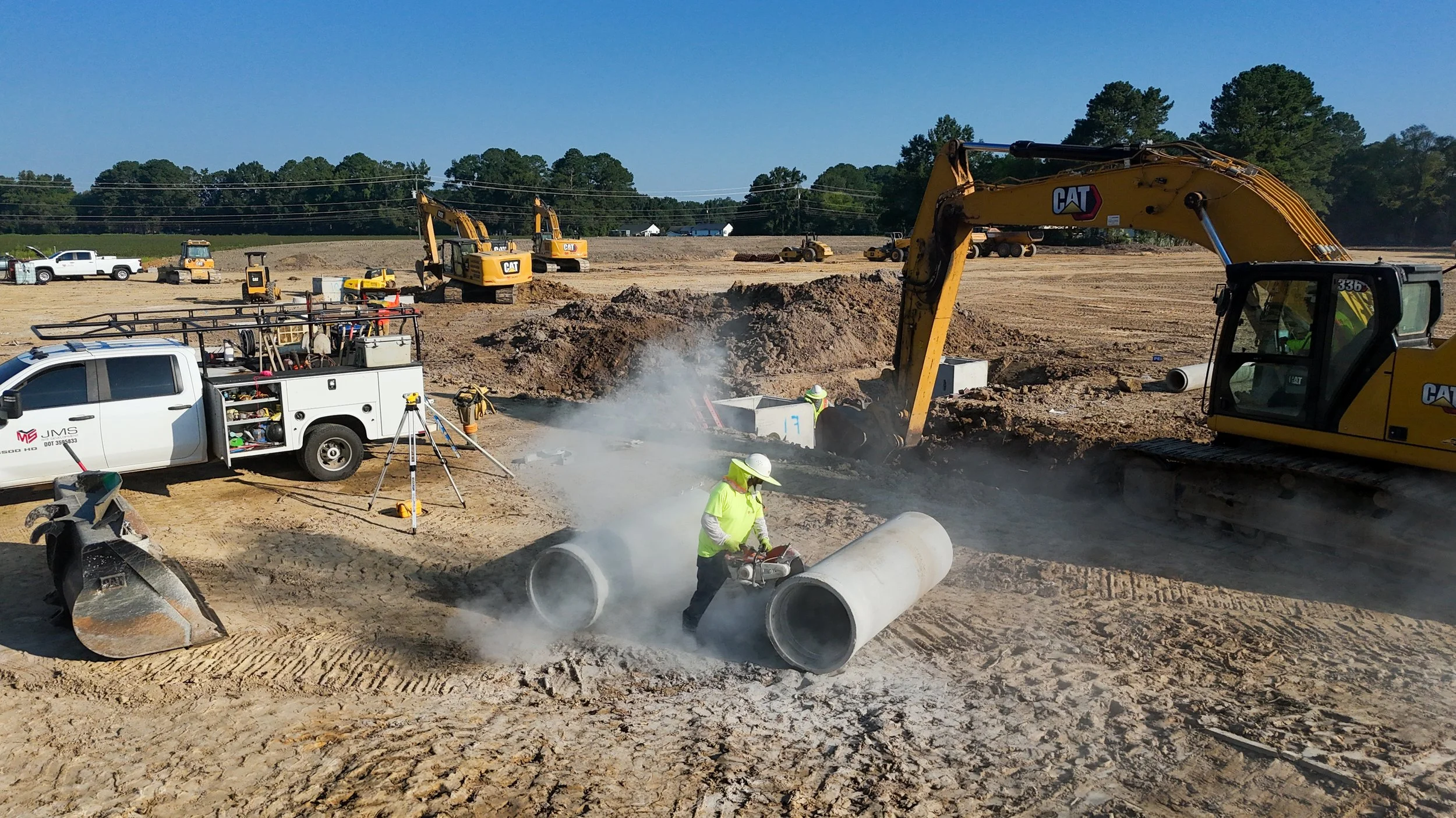 Construction site with multiple excavators, workers, and construction trucks working on underground pipe installation in a rural area with trees in the background.