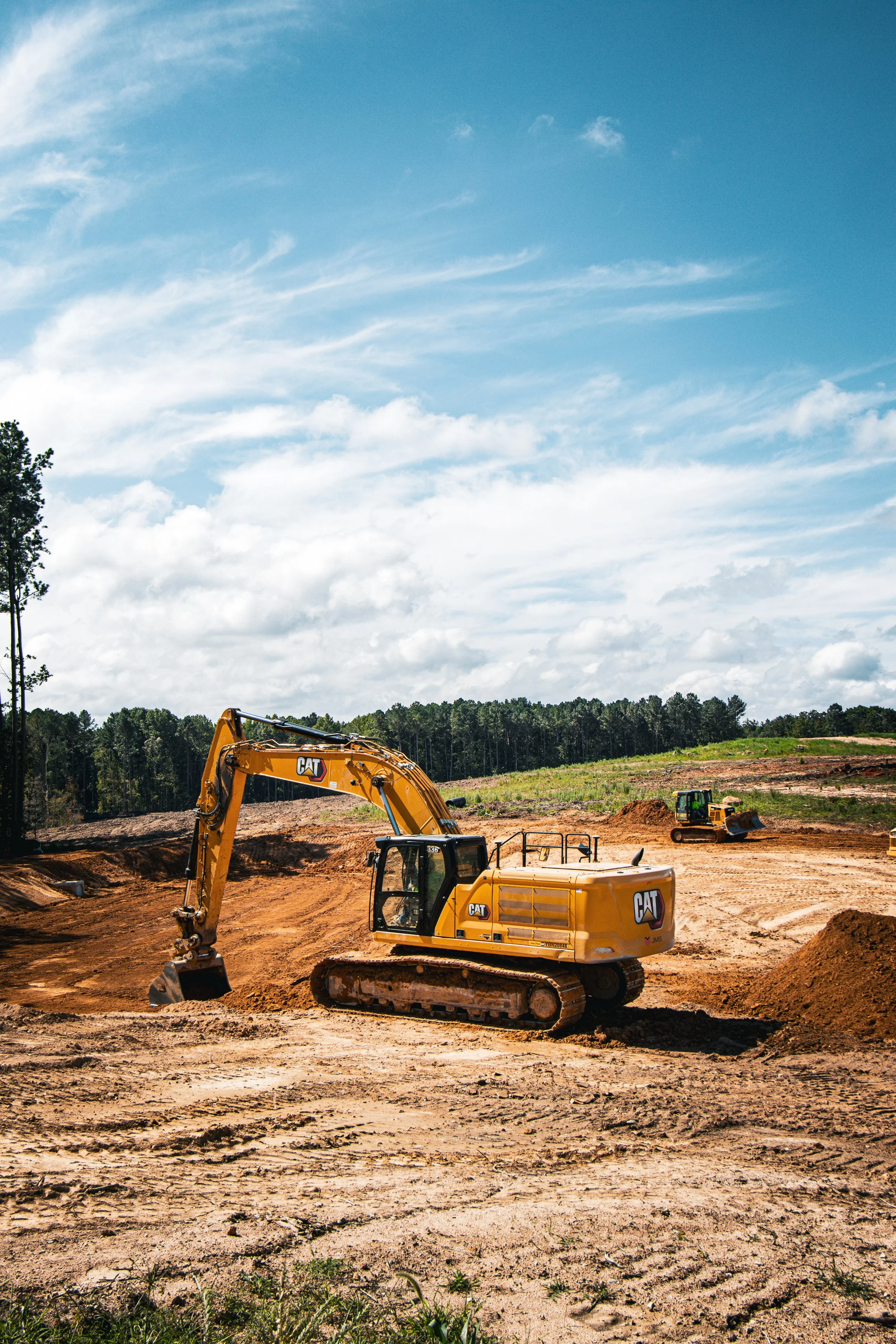 Construction site with a yellow excavator digging in red dirt under partly cloudy sky, with green trees in the background.