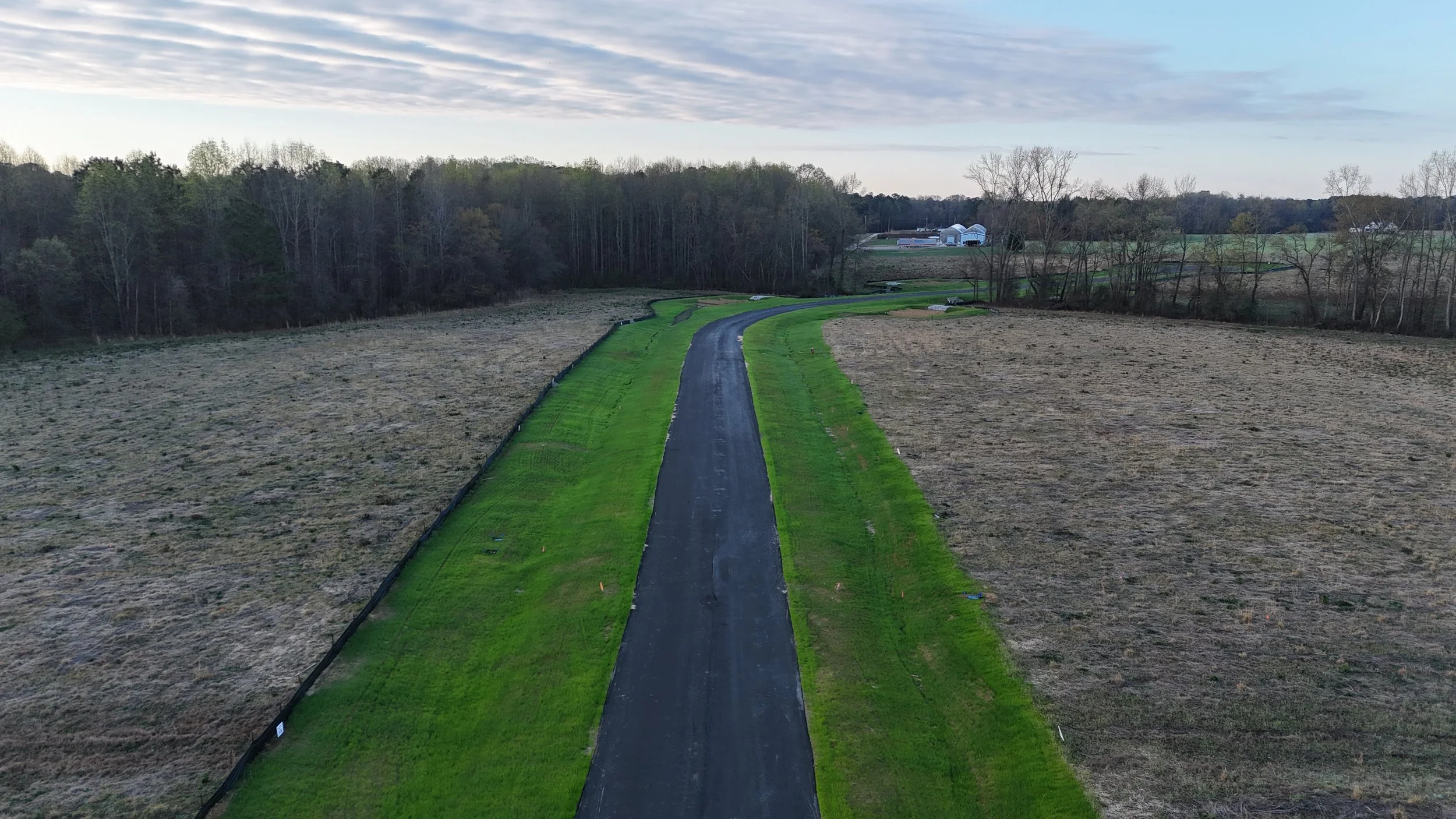 A winding paved road runs through a rural landscape with fields on either side. There are trees in the background and a few white buildings or greenhouses on the horizon under a partly cloudy sky.