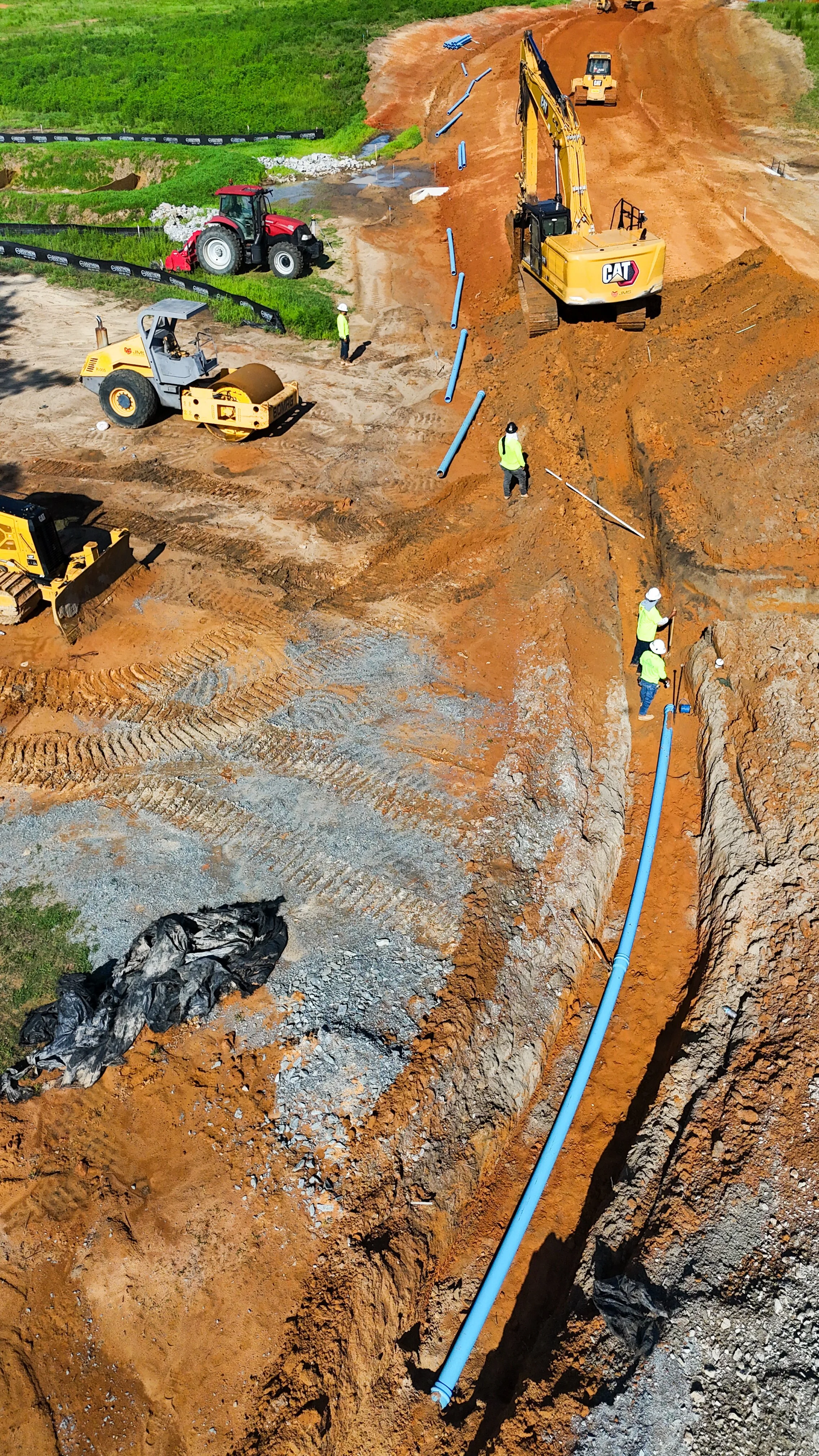 Construction workers installing underground piping on a construction site, with heavy machinery, dirt, and construction materials visible.