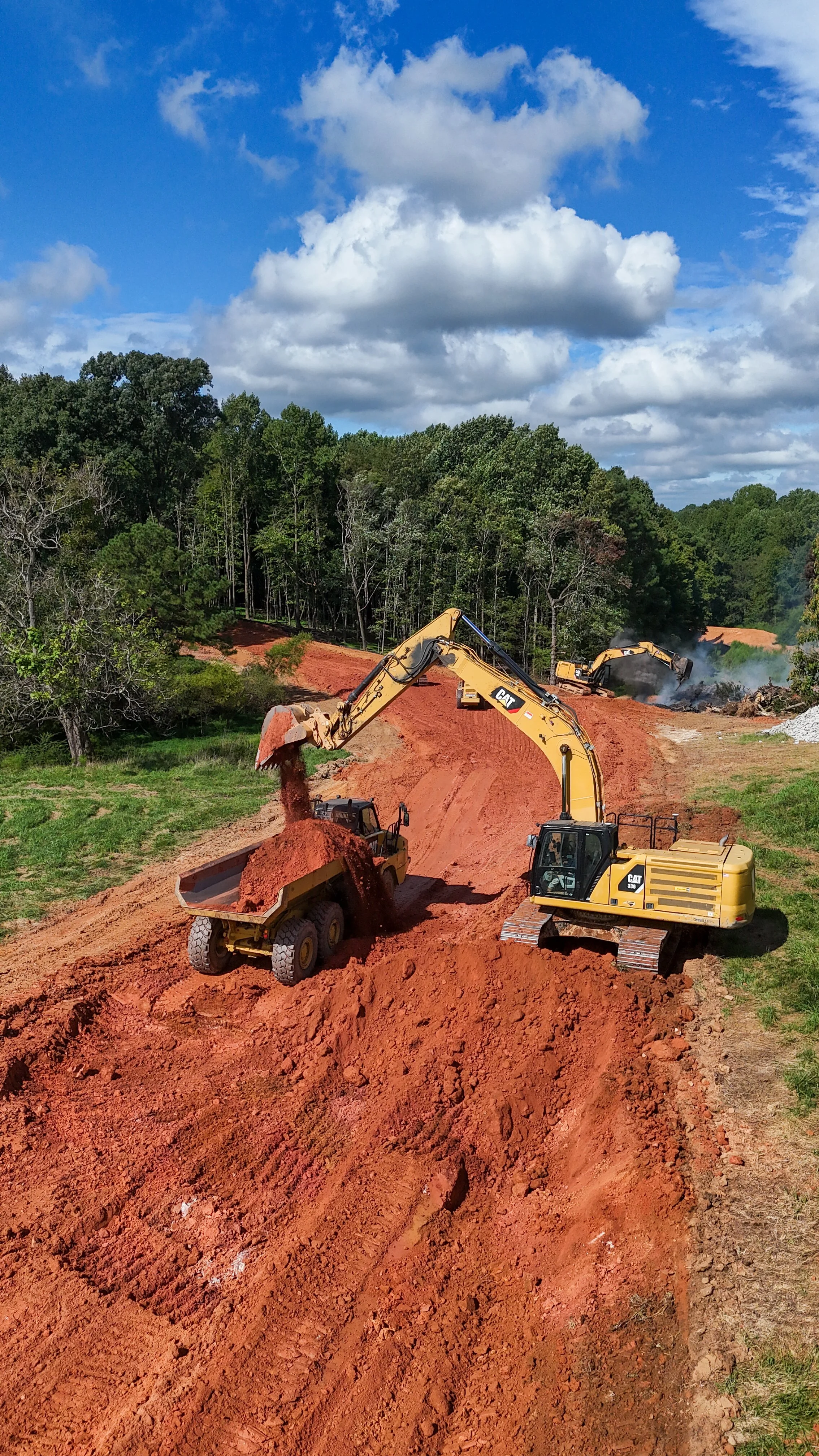 Construction site with two yellow excavators moving red dirt along a dirt road surrounded by green trees and a cloudy blue sky.