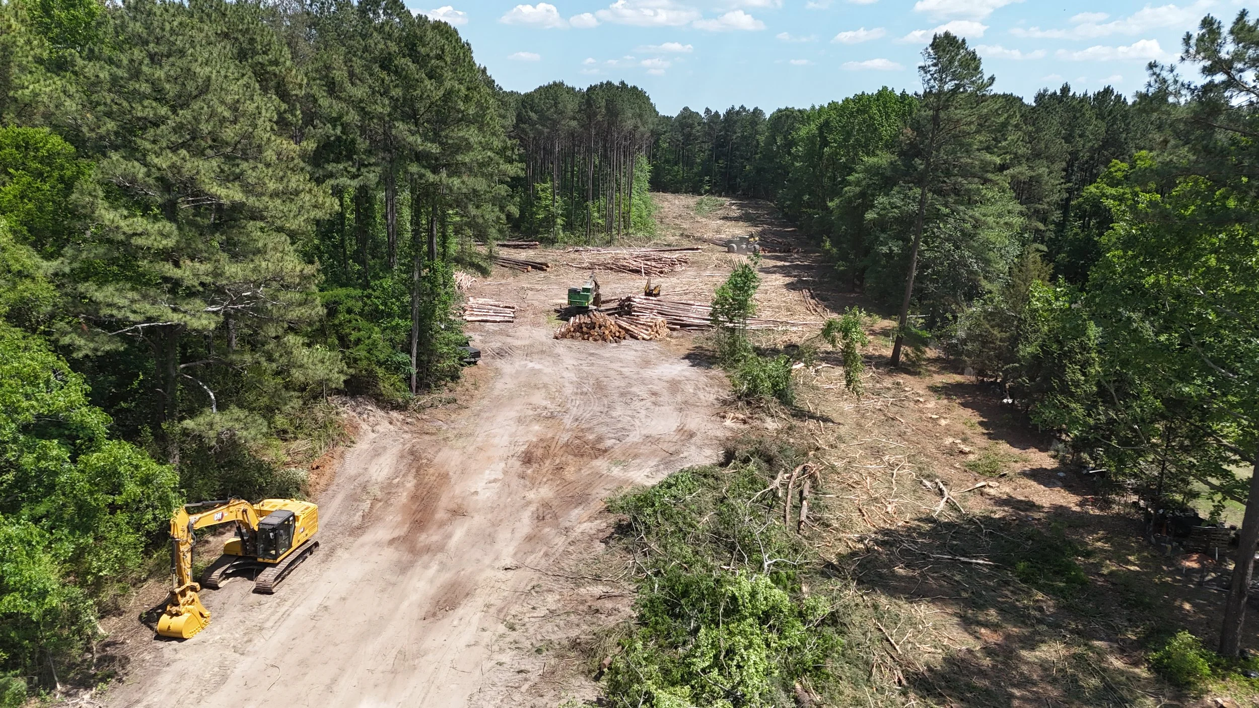 Aerial view of a forest clearing with logs and trees cut down, with construction machinery including an excavator and bulldozer on a dirt road.