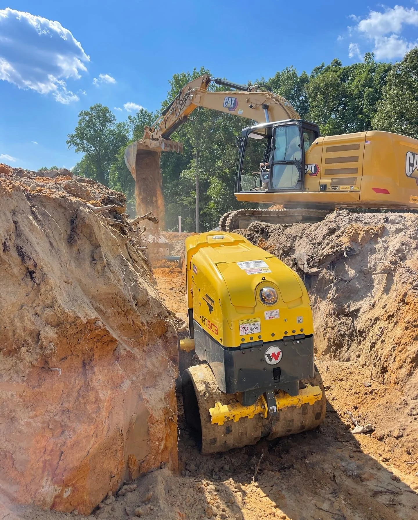 A bulldozer and an excavator working together at a construction site with dirt and rocks, under a blue sky with some clouds.