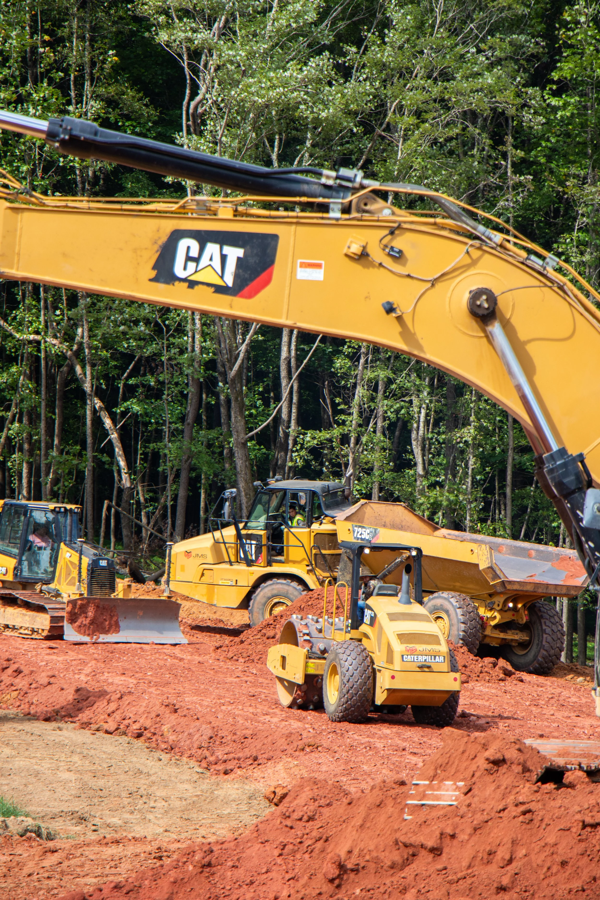 Construction site with multiple yellow Caterpillar heavy machinery working on earth movement and excavation, with a forested area in the background.