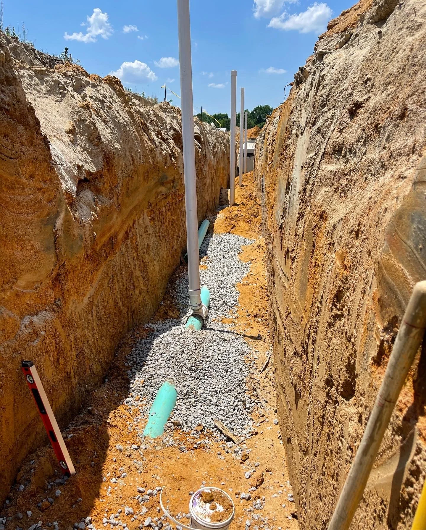 Underground construction trench with exposed orange soil, green pipes, and gravel, with vertical blue pipes and construction tools.