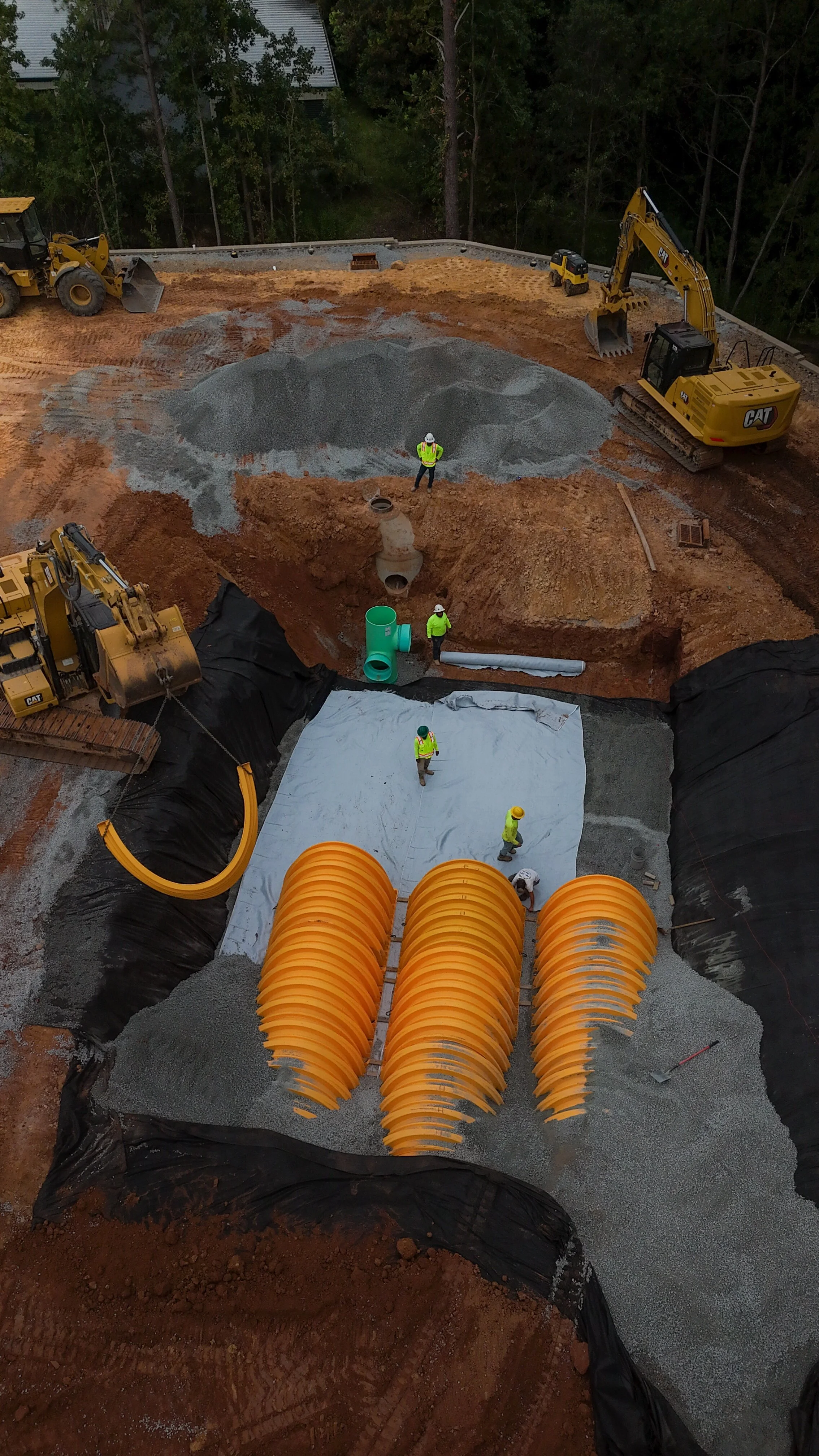 Construction site with workers and heavy machinery installing a large underground pipe system, including bright orange flexible pipes and gravel base.