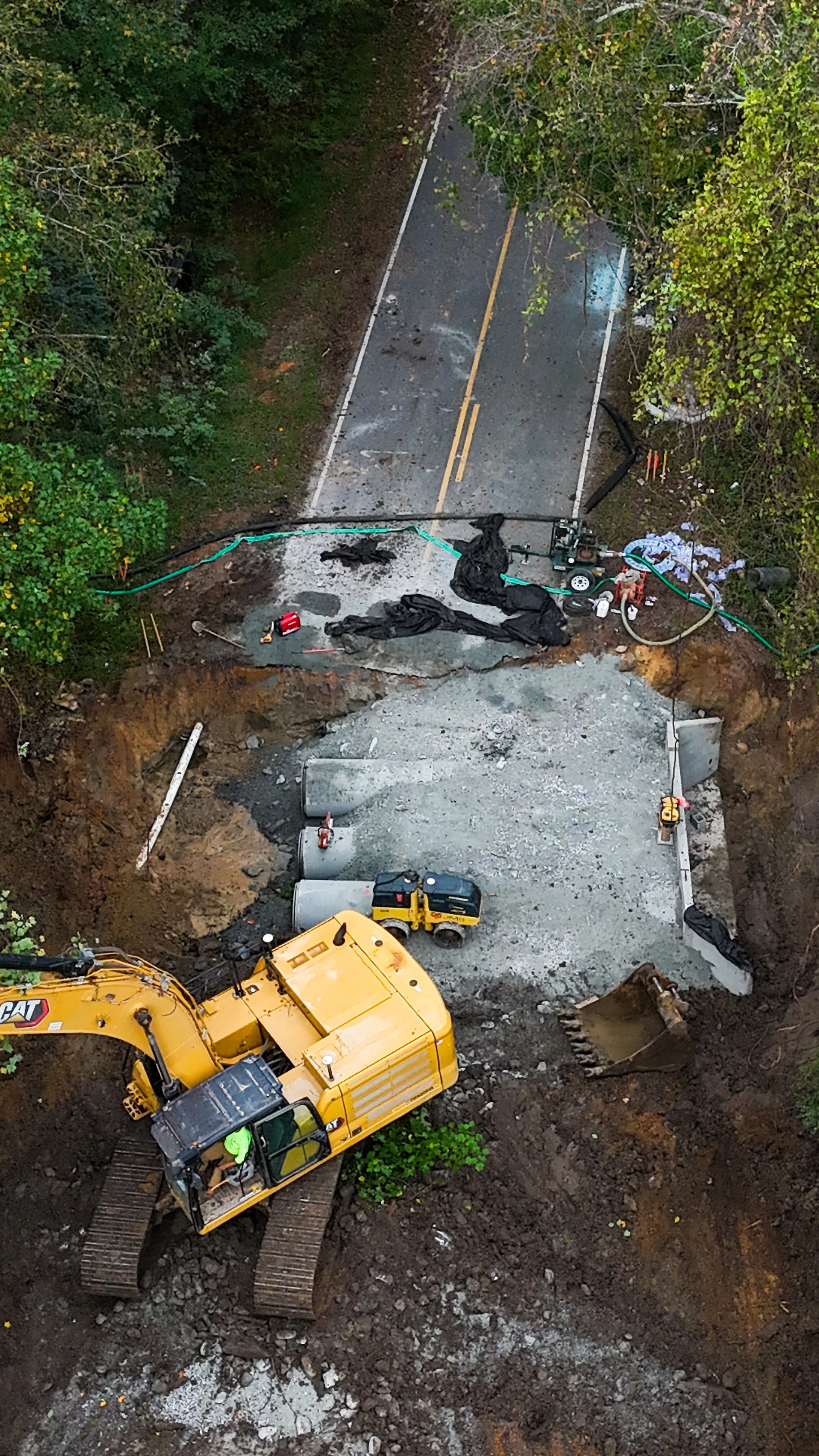 An aerial view of road construction site with a partially paved road, construction equipment like a yellow excavator and a small yellow construction vehicle, and equipment for laying concrete and fixing pipes.