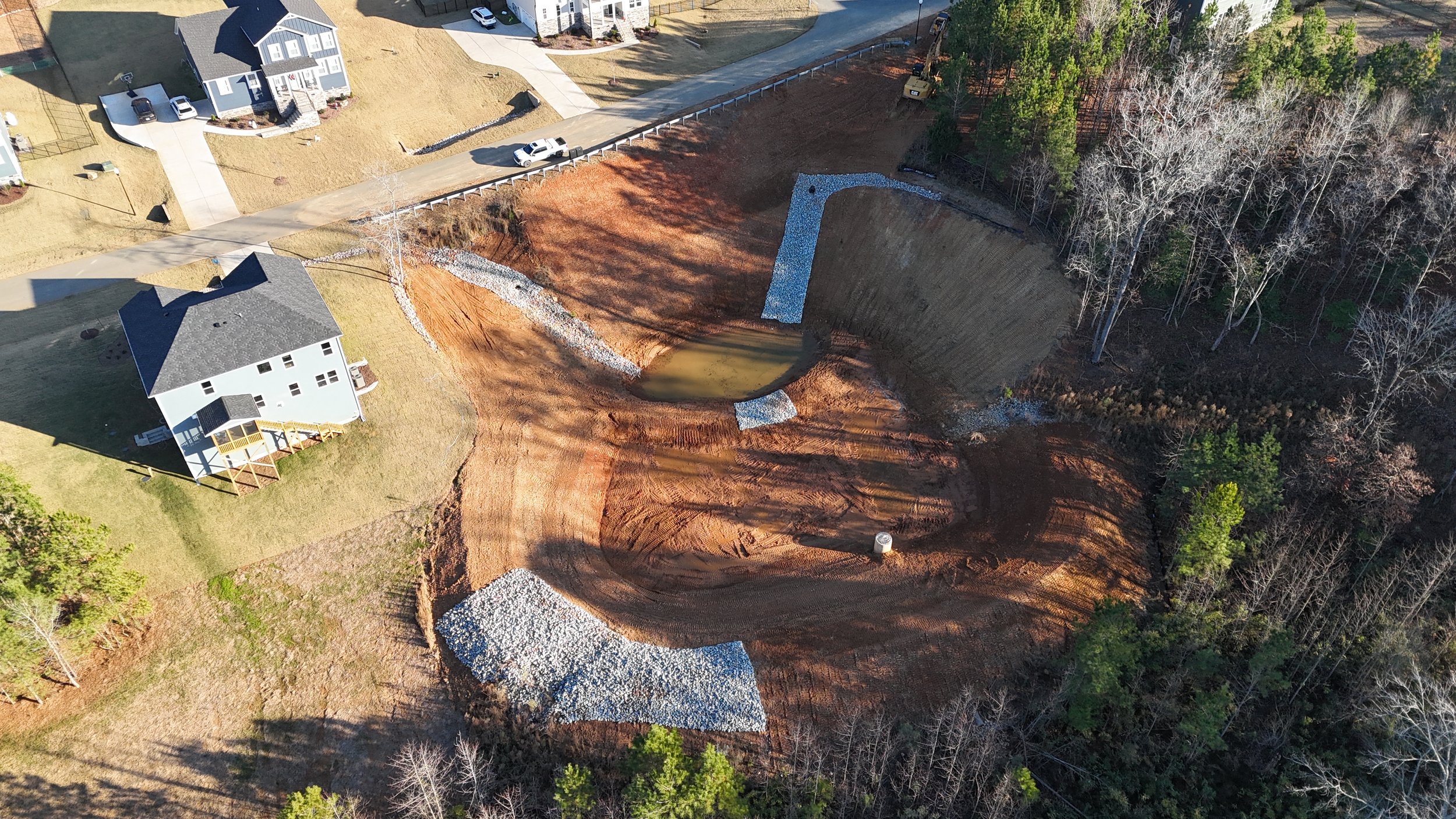 Aerial view of a residential area showing a house, nearby cars, and ongoing construction of an underground swimming pool with earth excavated around it, surrounded by trees and grassy yards.