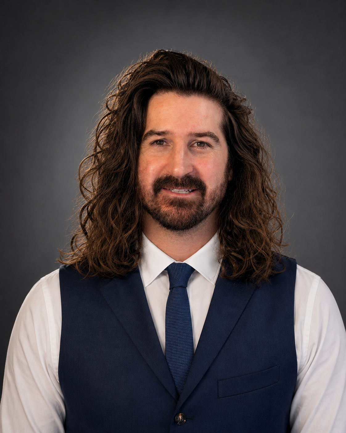 A man with long, curly brown hair and a beard, wearing a white shirt, a dark blue vest, and a matching tie, smiling against a dark gray background.