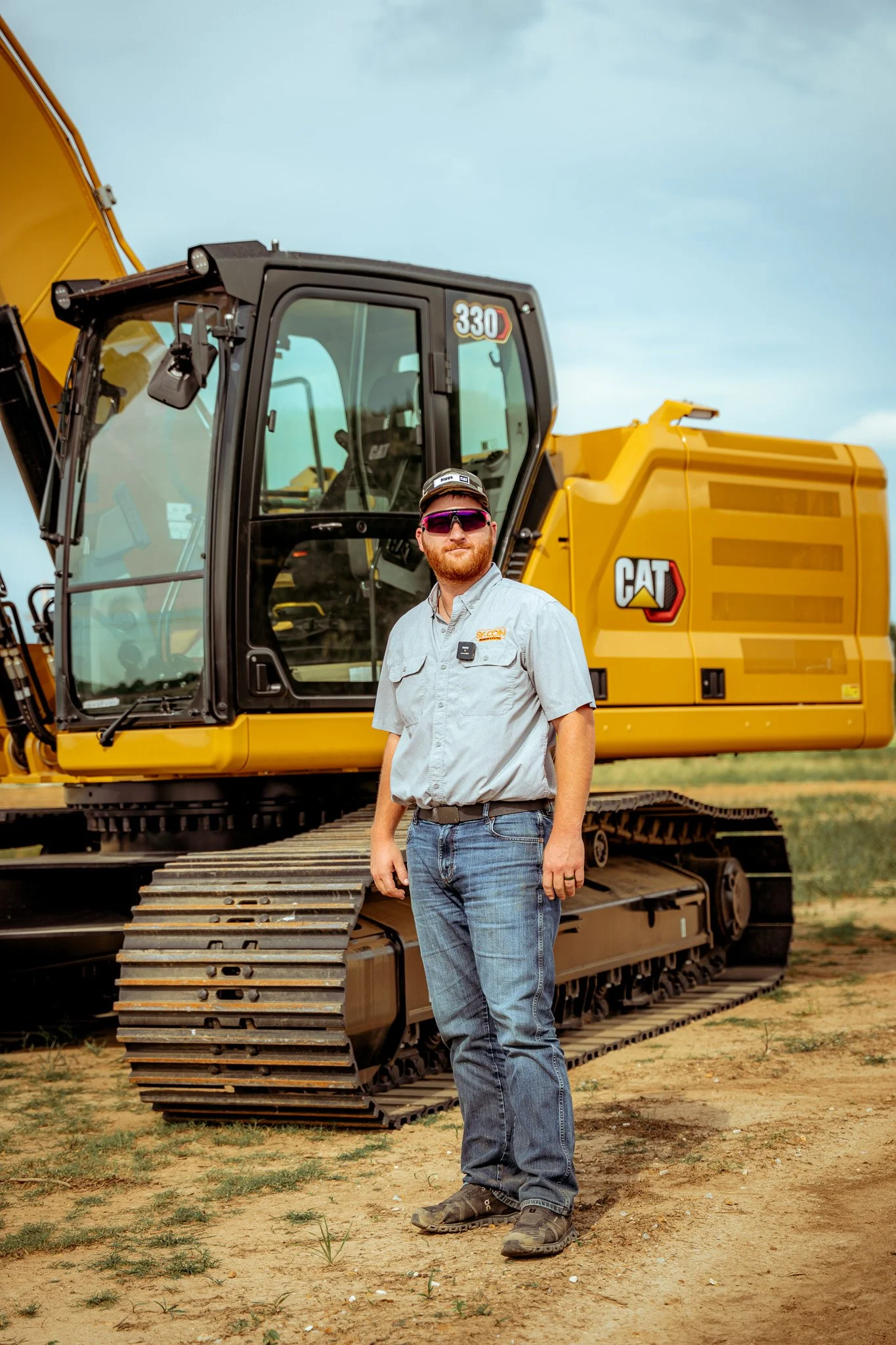 A man stands in front of a large yellow Caterpillar excavator on a dirt field with patches of grass, under a partly cloudy sky.