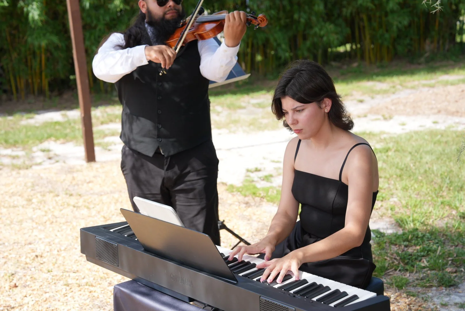 A woman playing a Yamaha digital piano outdoors, while a man with a violin stands behind her playing the instrument.