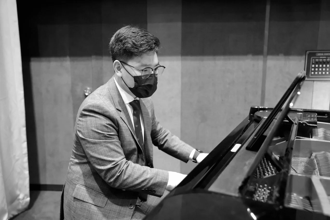 A man in a suit and glasses wearing a face mask playing a grand piano in a room with wood-paneled walls.
