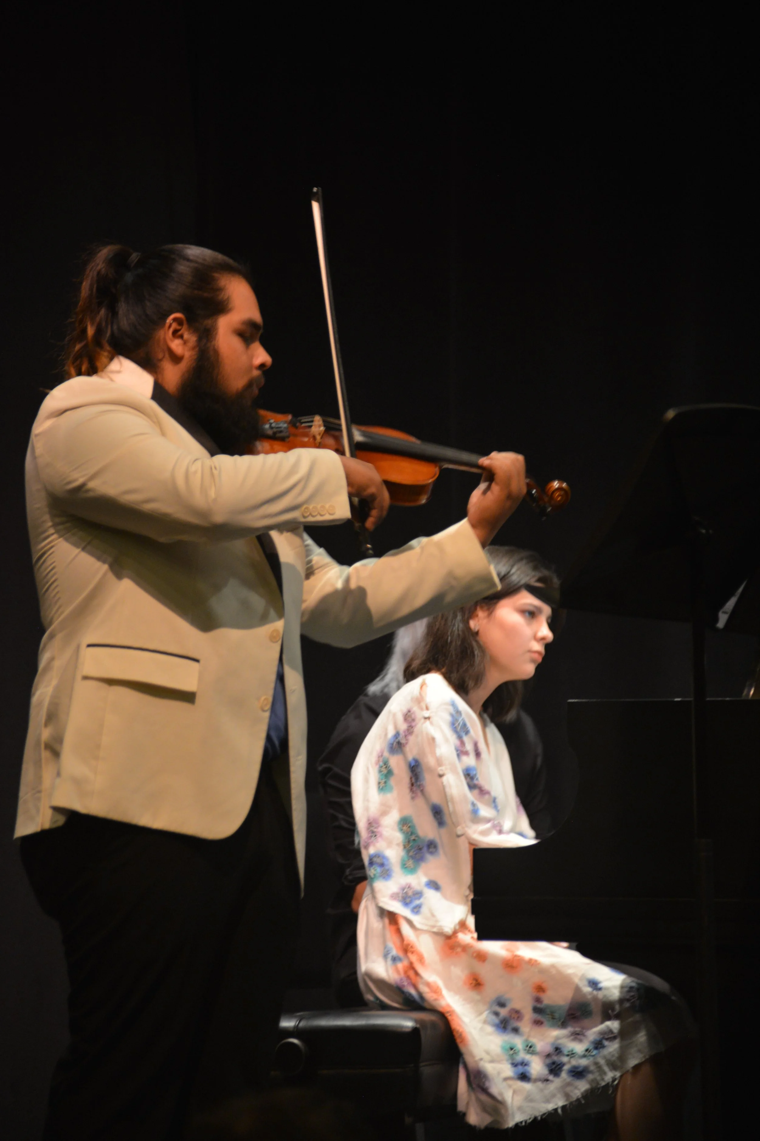 A man with a beard and long hair pulled back, dressed in a beige blazer, playing the violin. Sitting in front of a piano is a woman with shoulder-length brown hair wearing a floral dress, focused on the music.