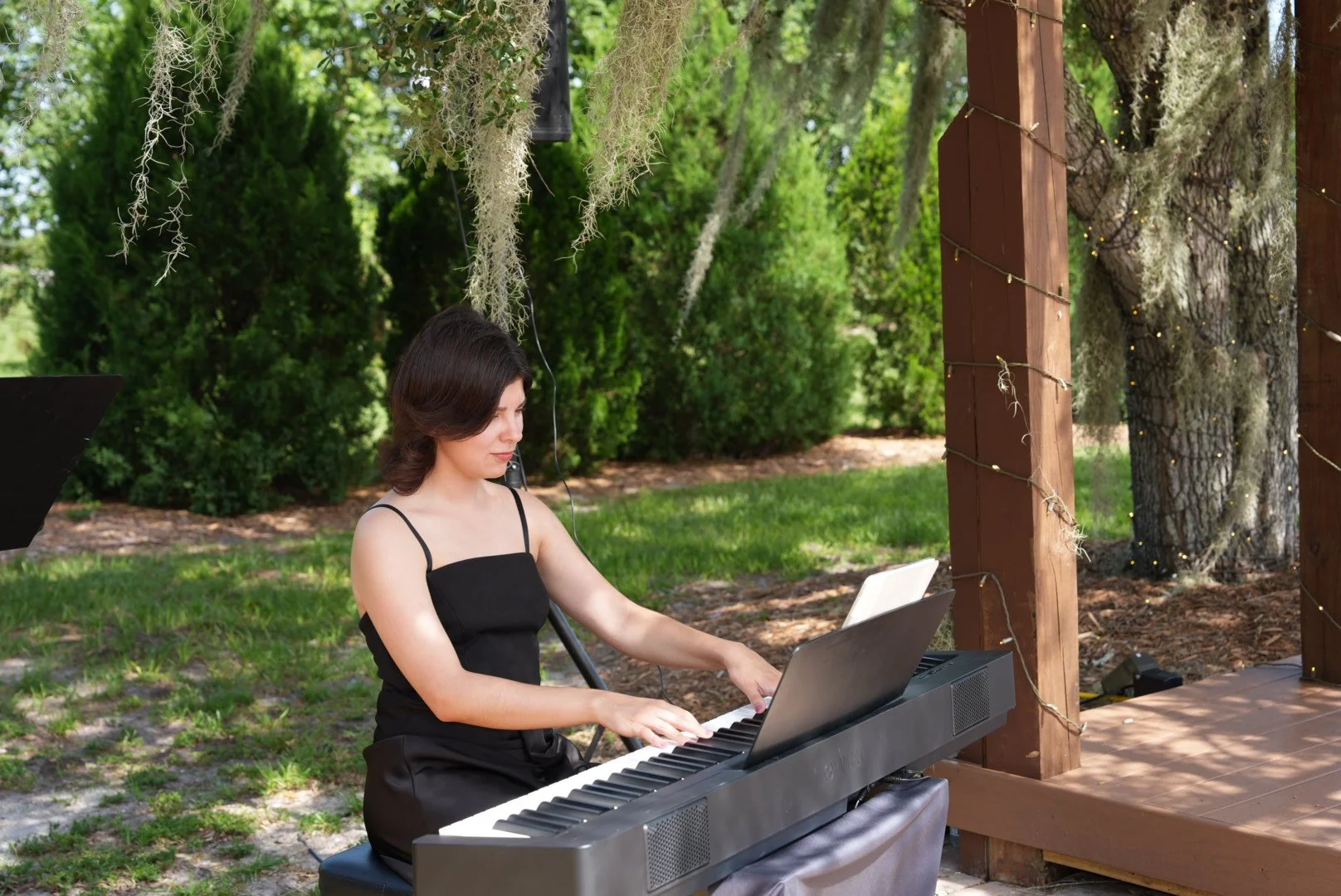 A woman in a black dress playing a digital piano outdoors on a wooden stage surrounded by trees and greenery.