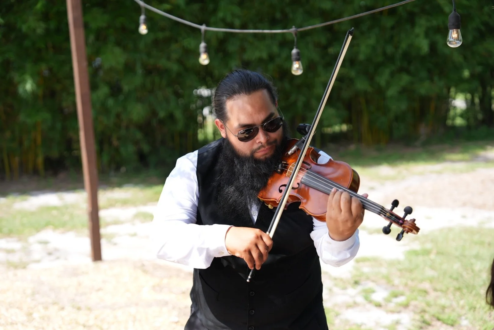 A man with a beard and long hair playing the violin outdoors in a garden surrounded by green trees and plants, using a smartphone mounted on a stand.