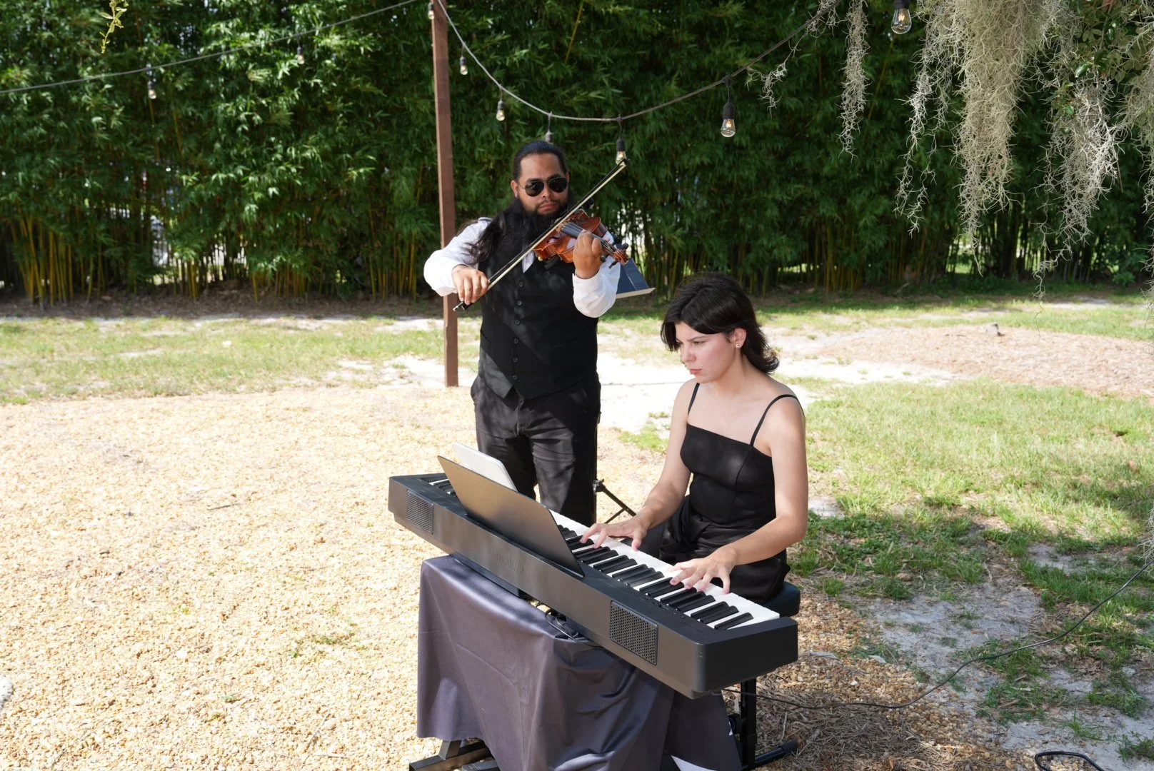 A woman playing a keyboard outdoors while a man plays violin behind her.