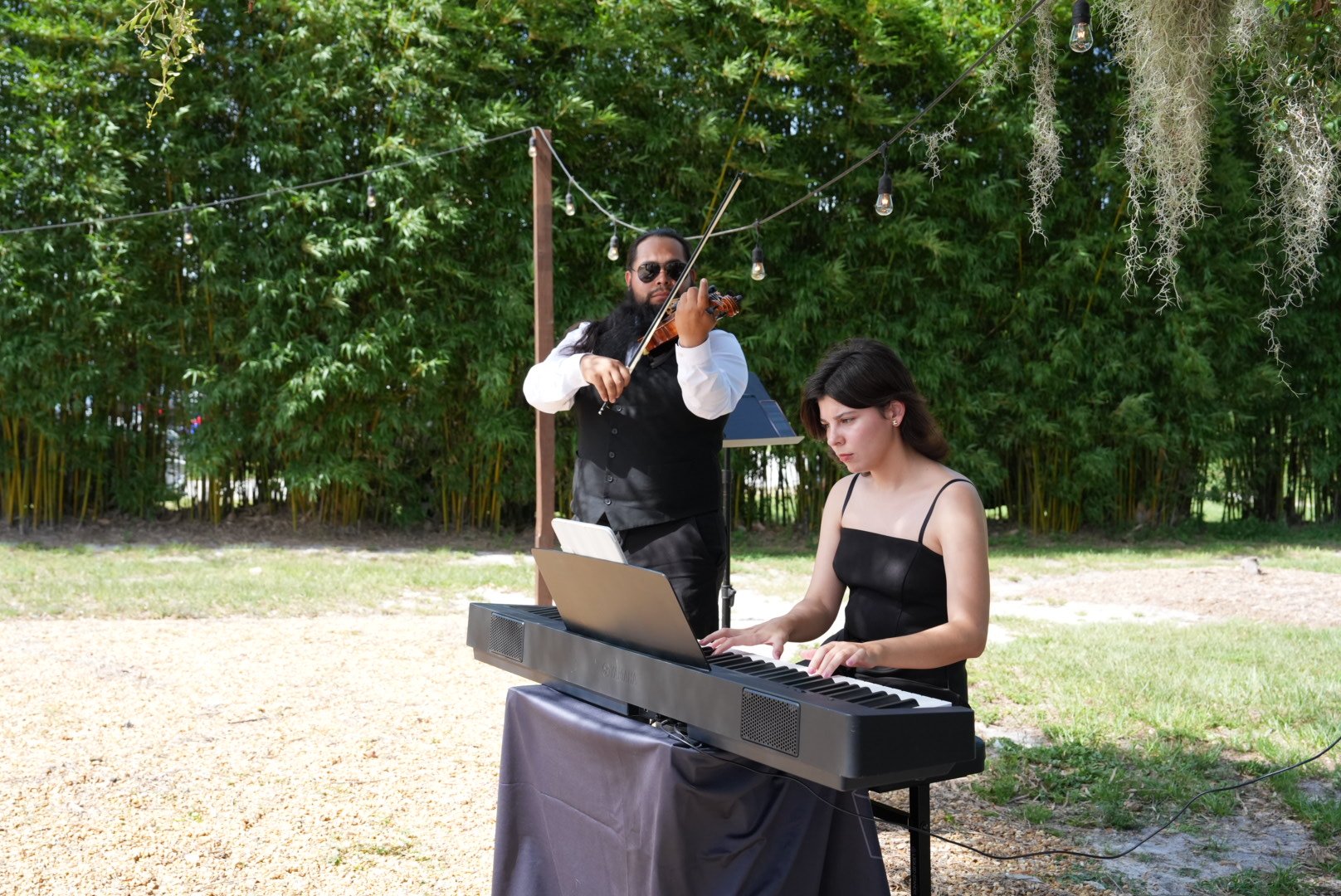 A woman playing a keyboard outdoors while a man playing a violin stands behind her. They are surrounded by greenery and string lights hang above them.