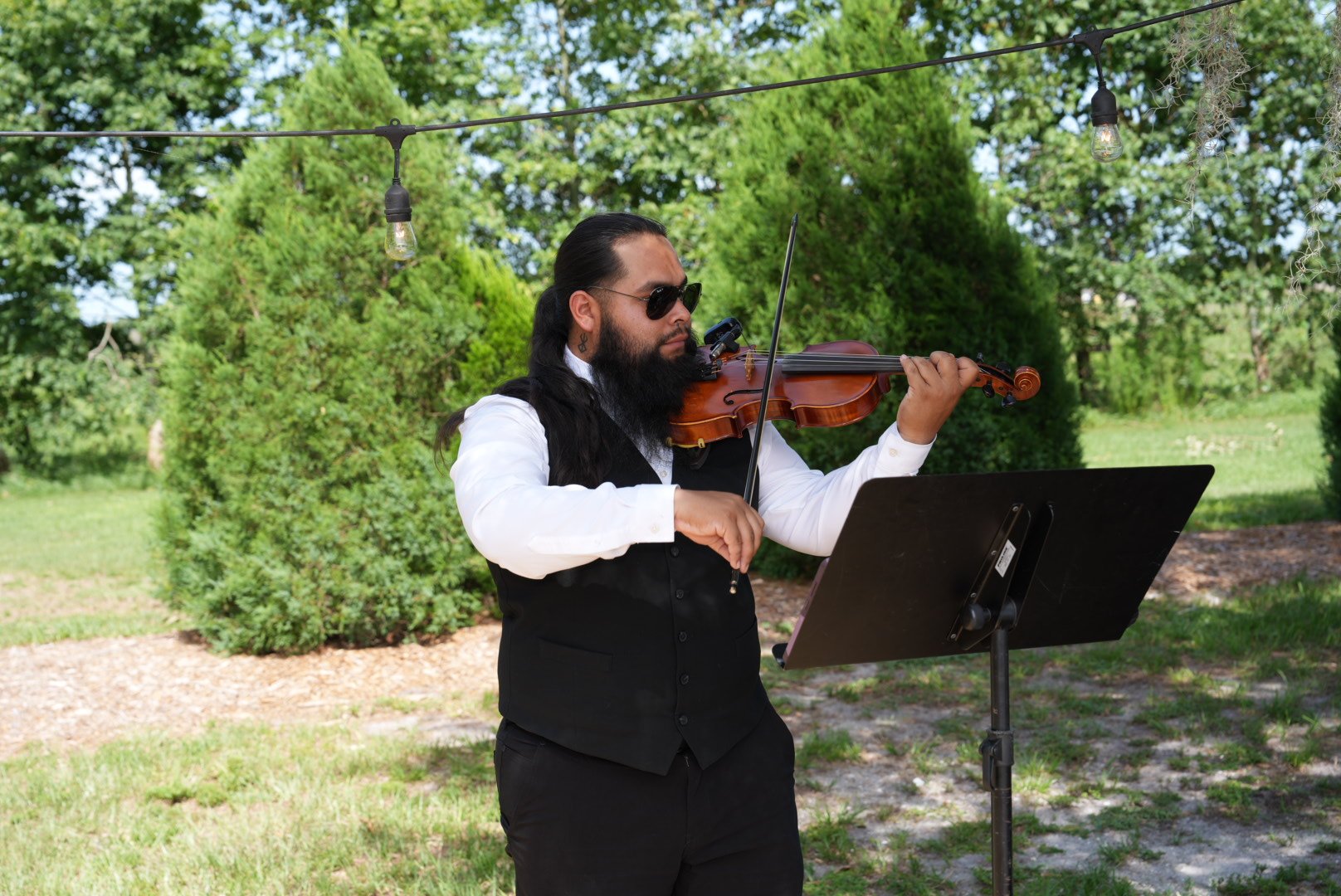 A man with long black hair, a beard, sunglasses, and a white shirt playing a violin outdoors with trees and string lights in the background.