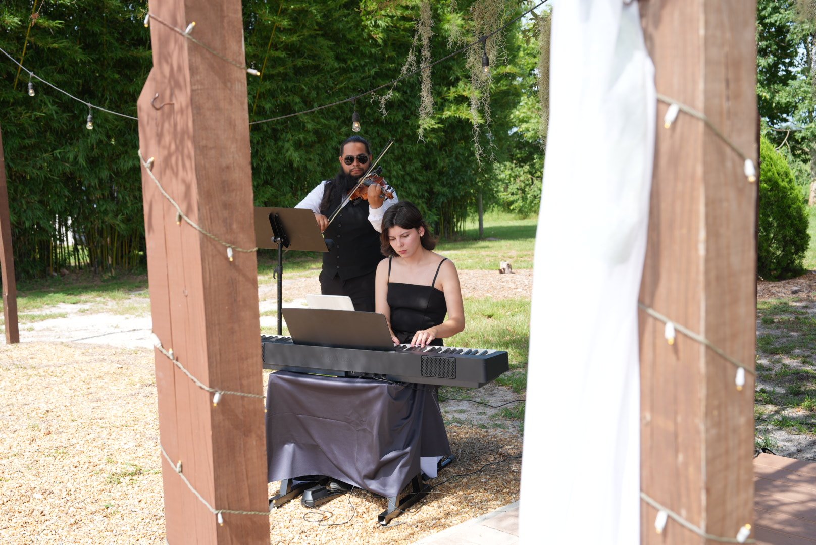 A woman playing a keyboard and a man playing a violin performing outdoors during daytime, framed by wooden posts and string lights.