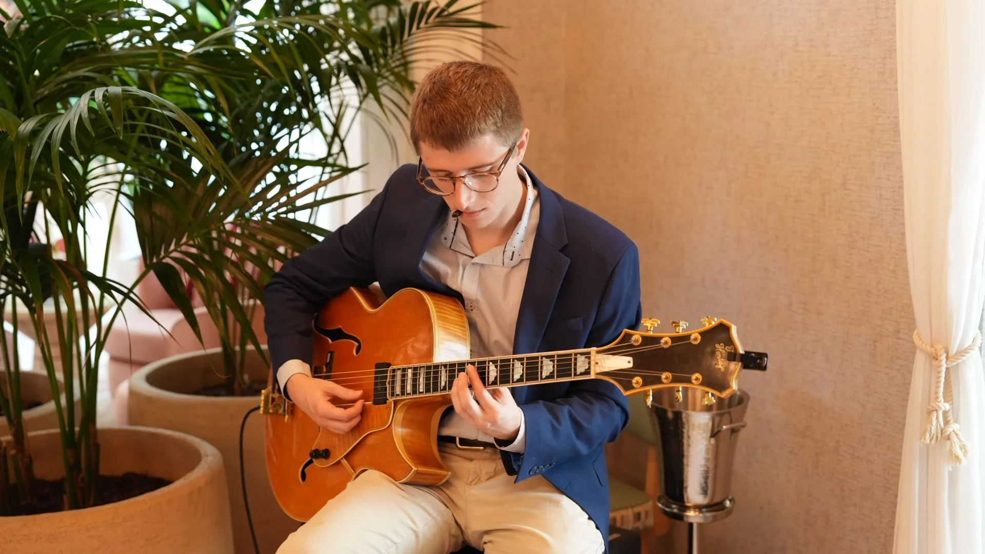 A young man with glasses playing an acoustic guitar indoors, surrounded by potted plants and beige curtains.