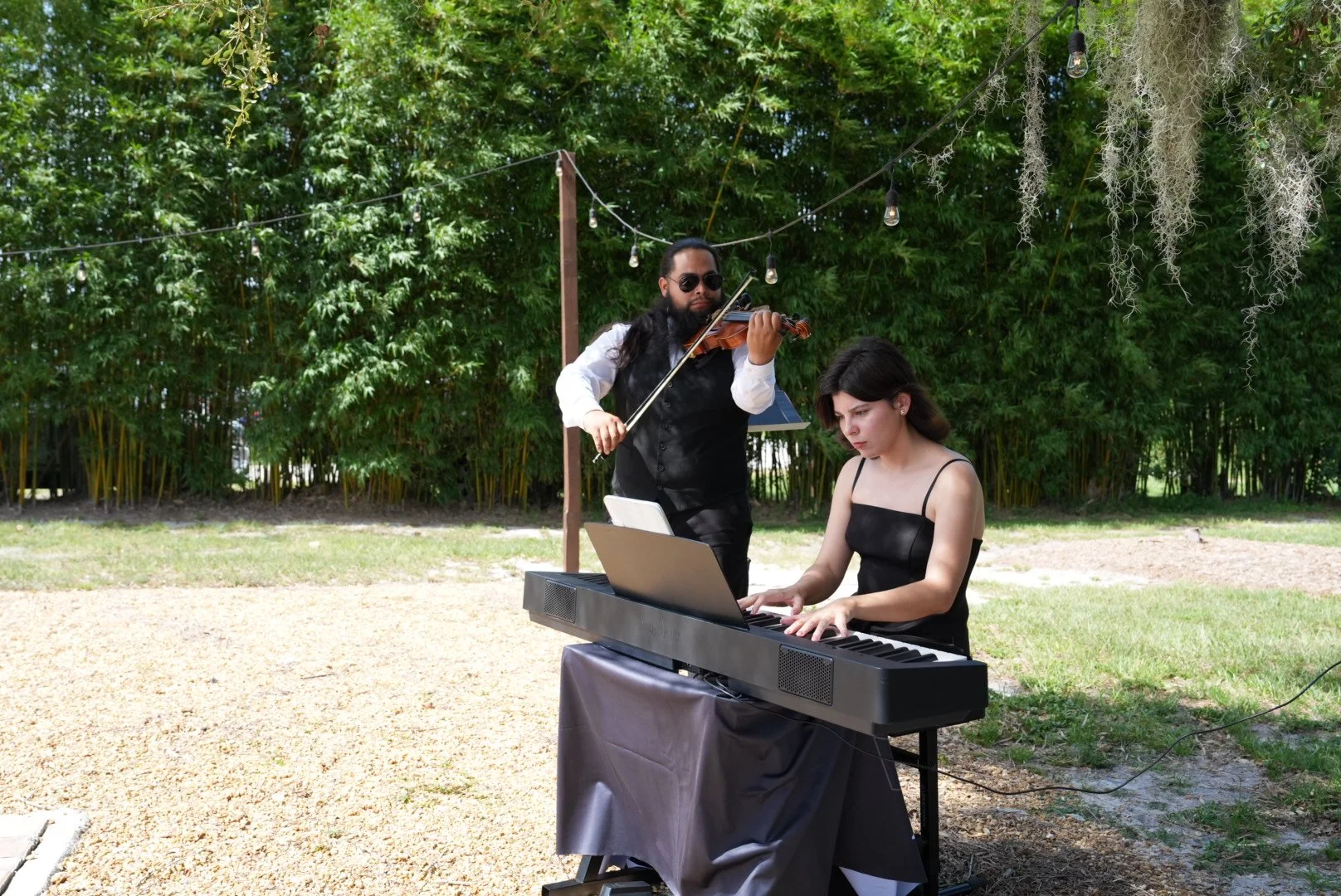 A woman playing the piano outdoors with a man playing the violin beside her. They are surrounded by green trees and string lights hanging overhead.