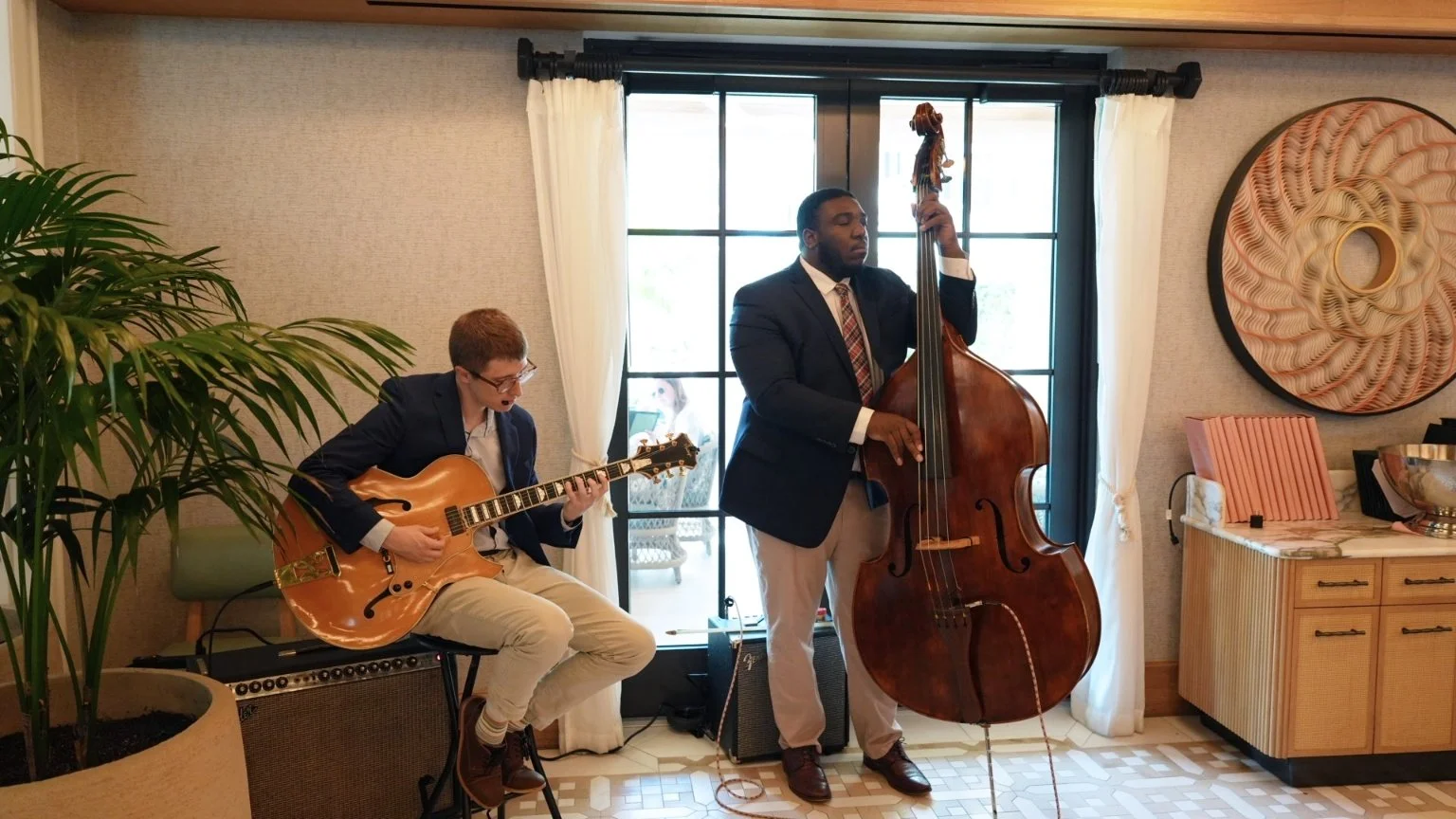 Two musicians playing jazz in a cozy indoor setting. One is sitting on a green chair, playing an acoustic guitar, and the other is standing, playing a double bass. They are dressed in formal attire with blazers.