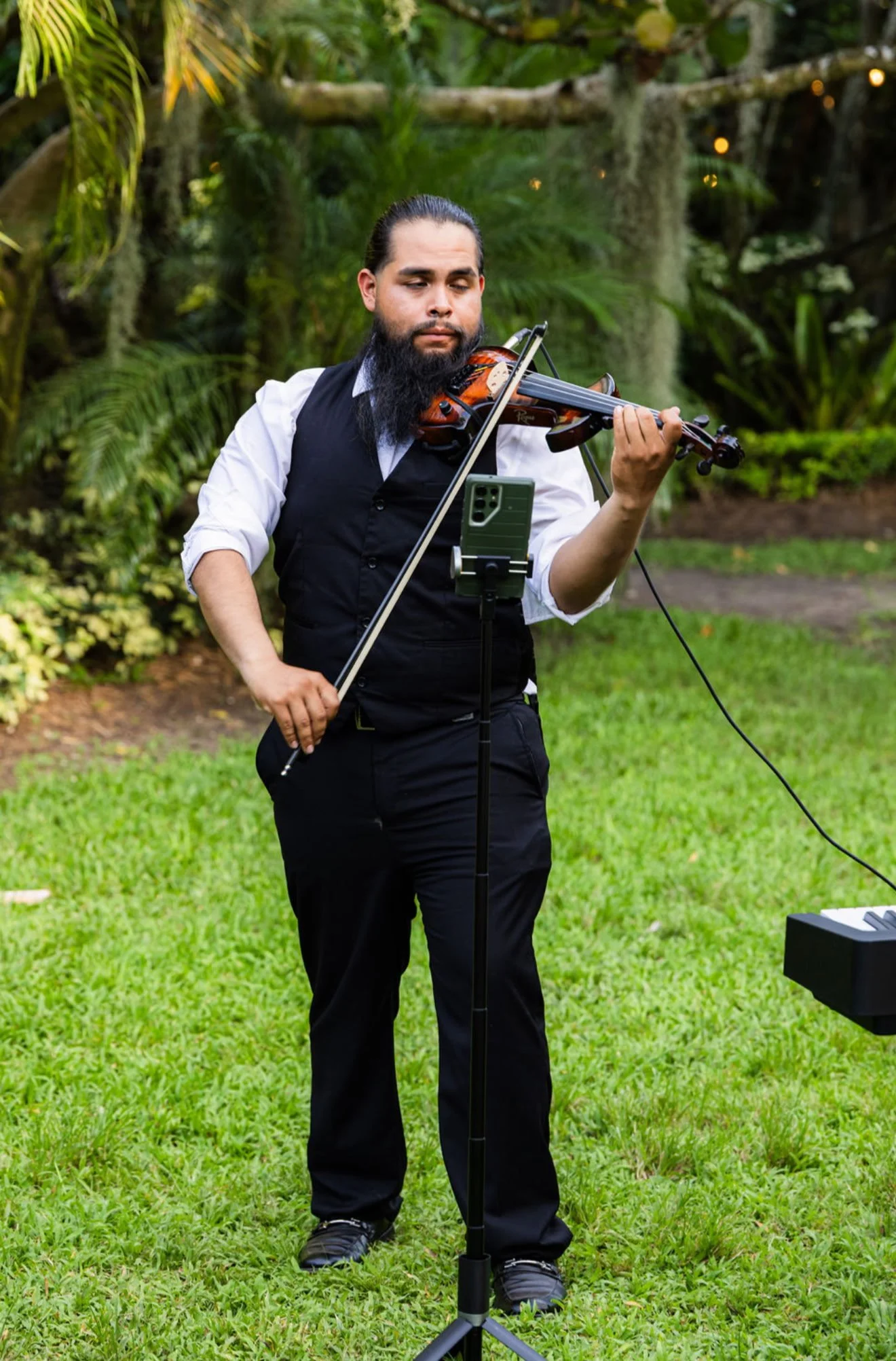 A man with a long beard and slicked-back hair wearing a white shirt, black vest, and black pants playing a violin outdoors. He stands on a grassy area with trees in the background and a music stand and electronic device in front of him.
