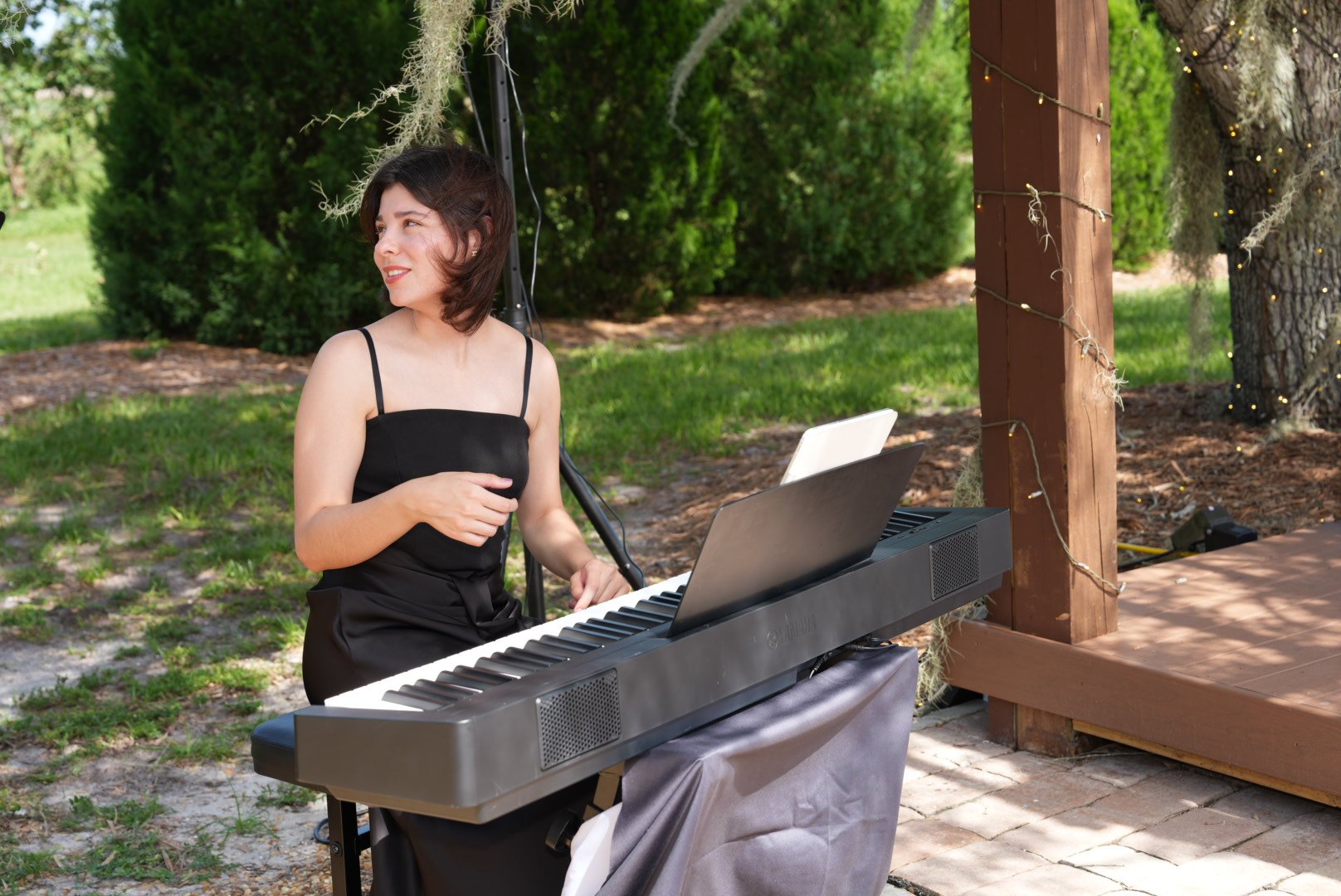 A woman in a black dress playing a digital keyboard outdoors under a wooden structure with string lights, surrounded by green trees and grass.