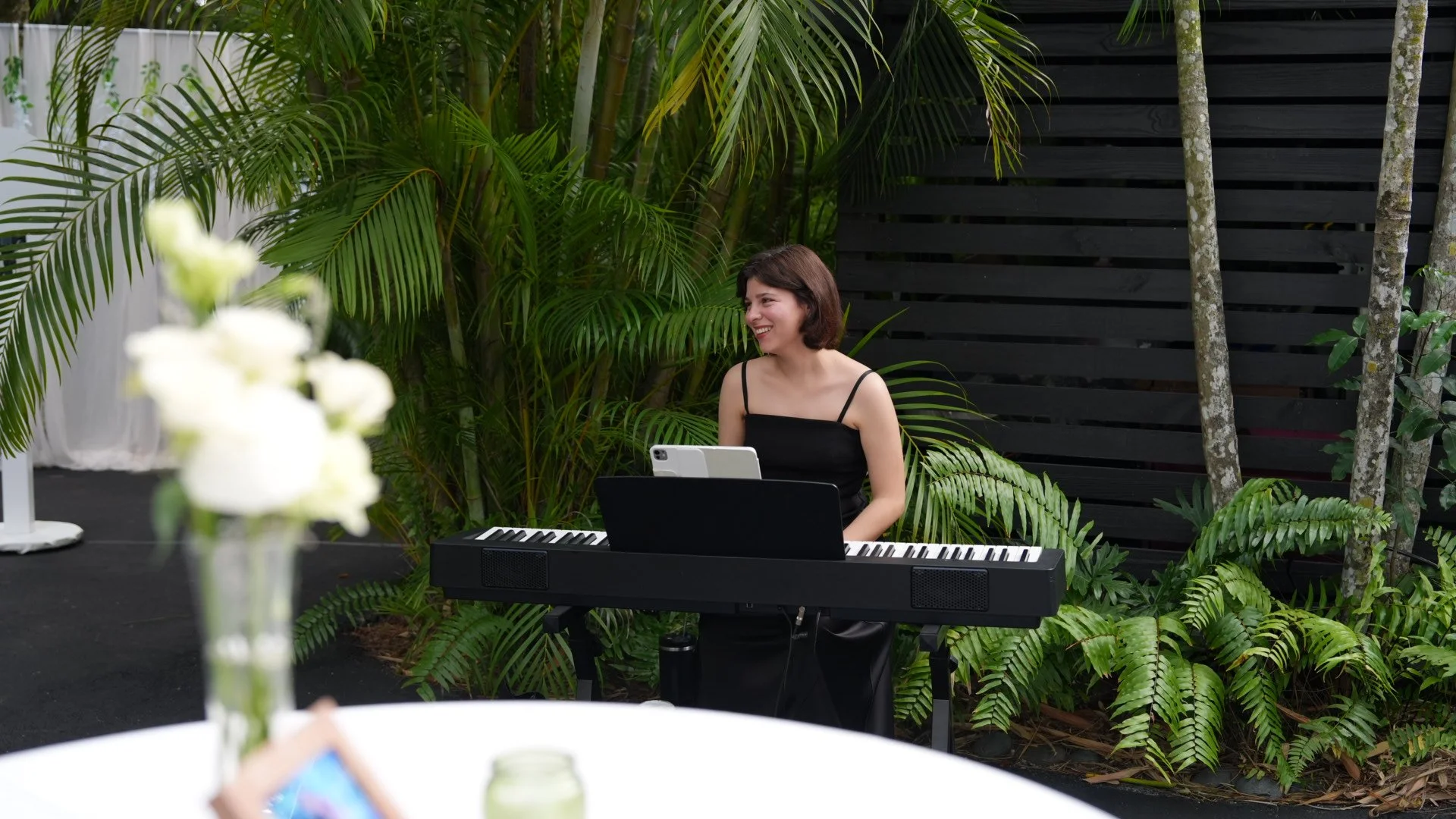 A woman sitting outdoors at a keyboard, smiling, surrounded by lush green plants and trees, with a black wooden fence in the background.