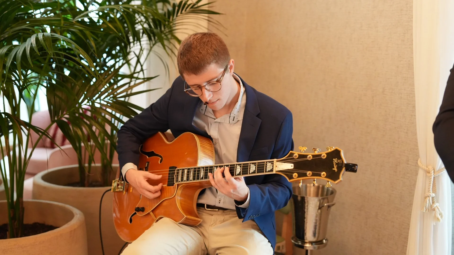 Young man with glasses playing an orange acoustic guitar, sitting indoors near a large potted plant and a pink couch.