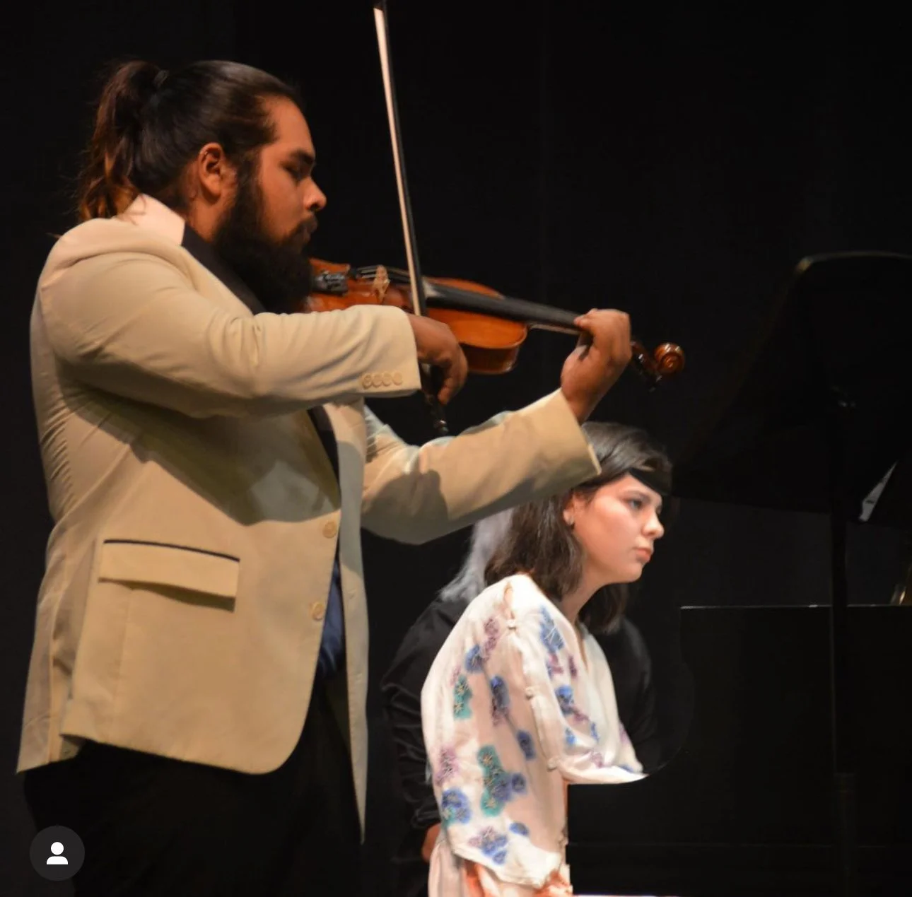 A man with long hair and a beard playing the violin while a woman sits at a piano, listening.