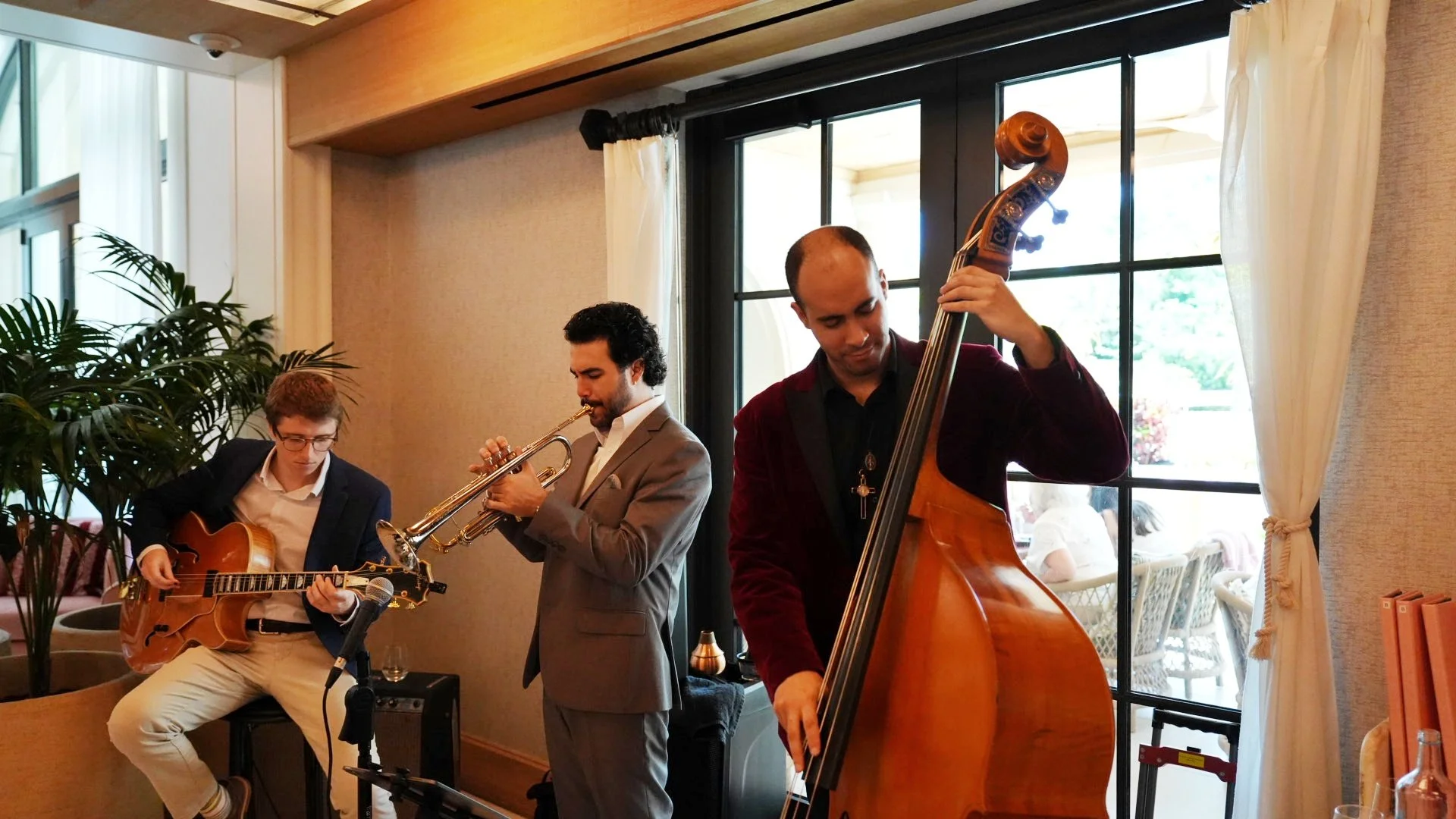 Three musicians performing indoors; a guitarist, a trumpet player, and a double bass player.