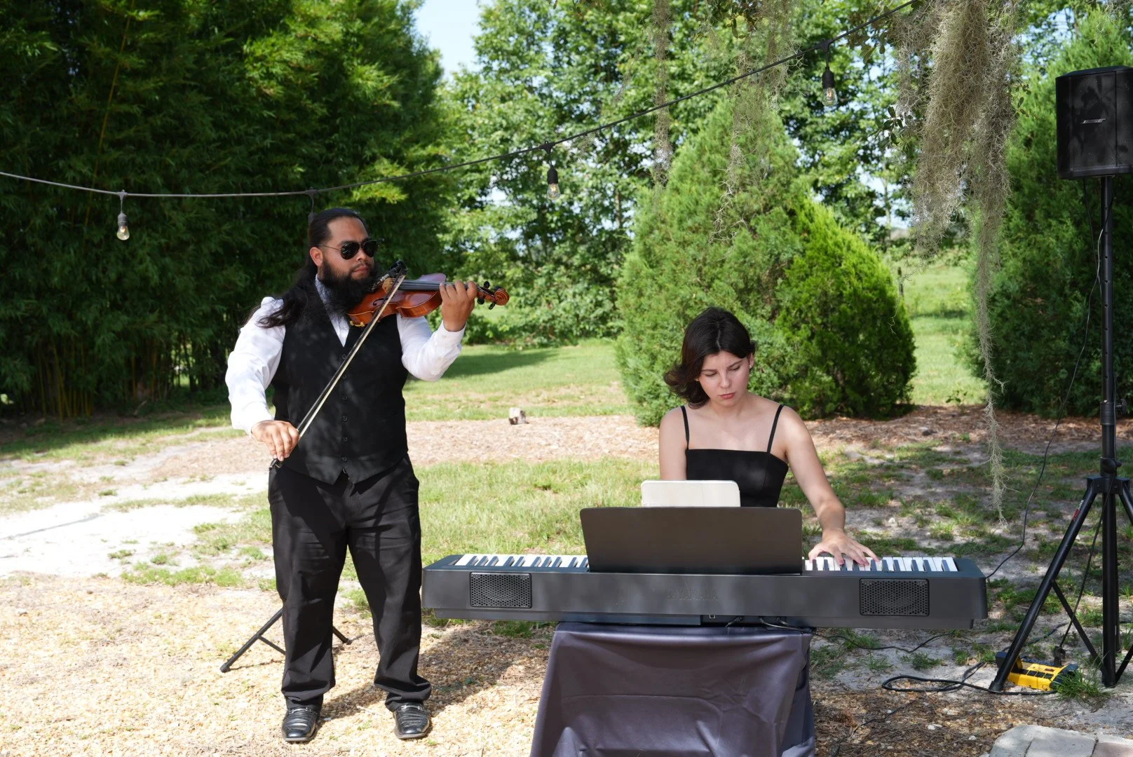 A man playing the violin while a woman is seated at an electronic keyboard outdoors under string lights, surrounded by green trees.