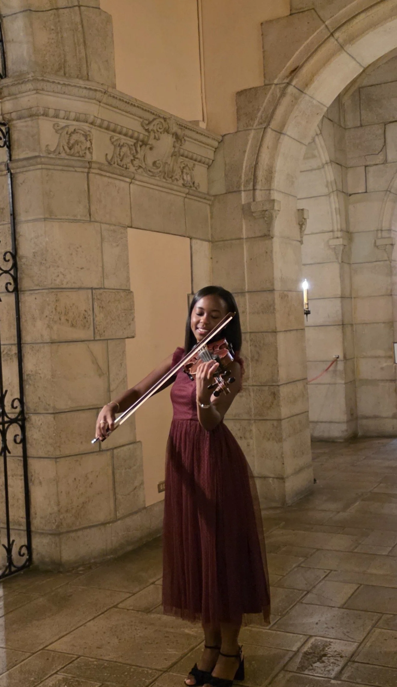 A woman in a long, burgundy dress playing a violin in a stone archway and hallway at night, illuminated by candlelight.