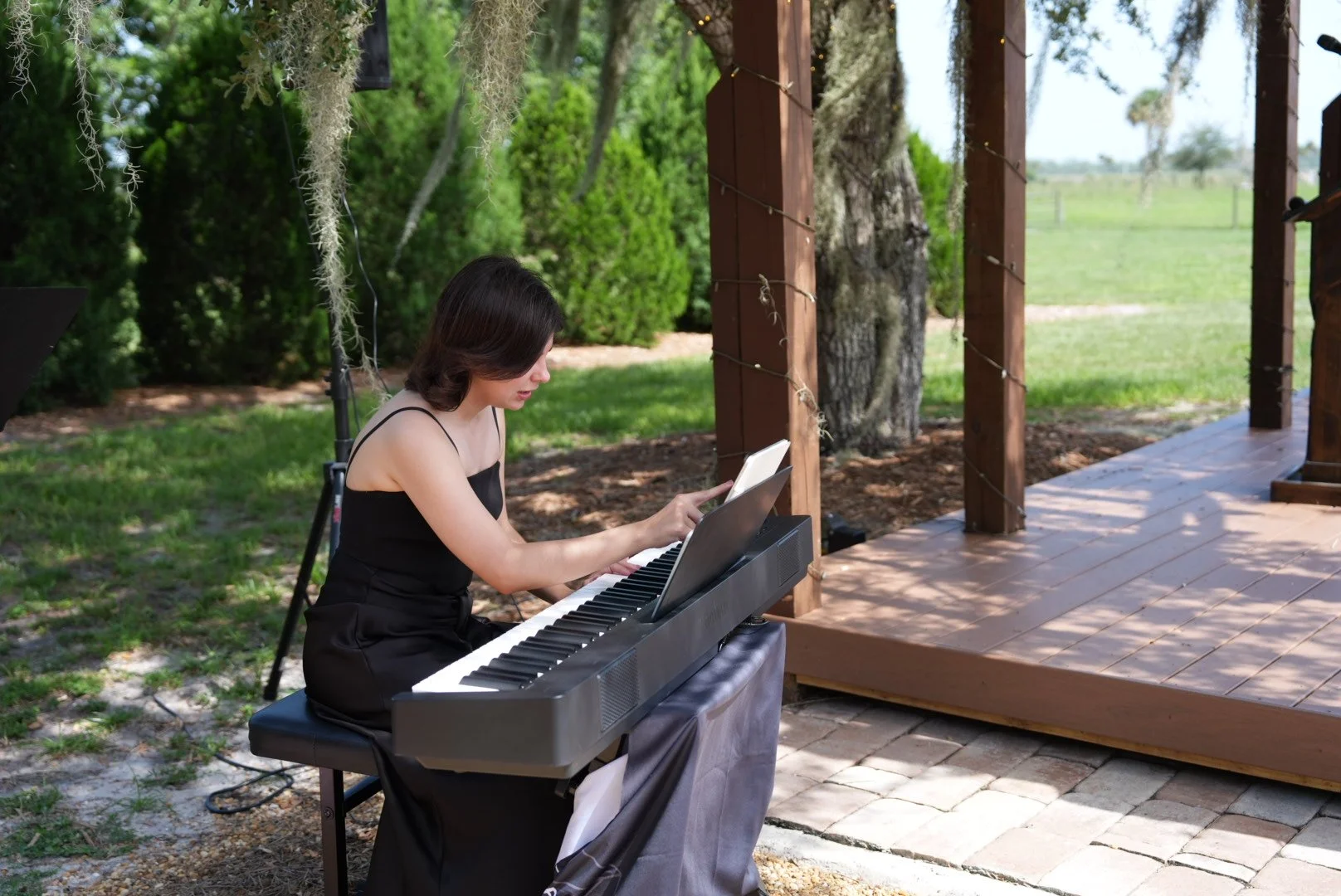 Woman in black dress playing digital piano outdoors under a shaded porch with trees and greenery in the background.