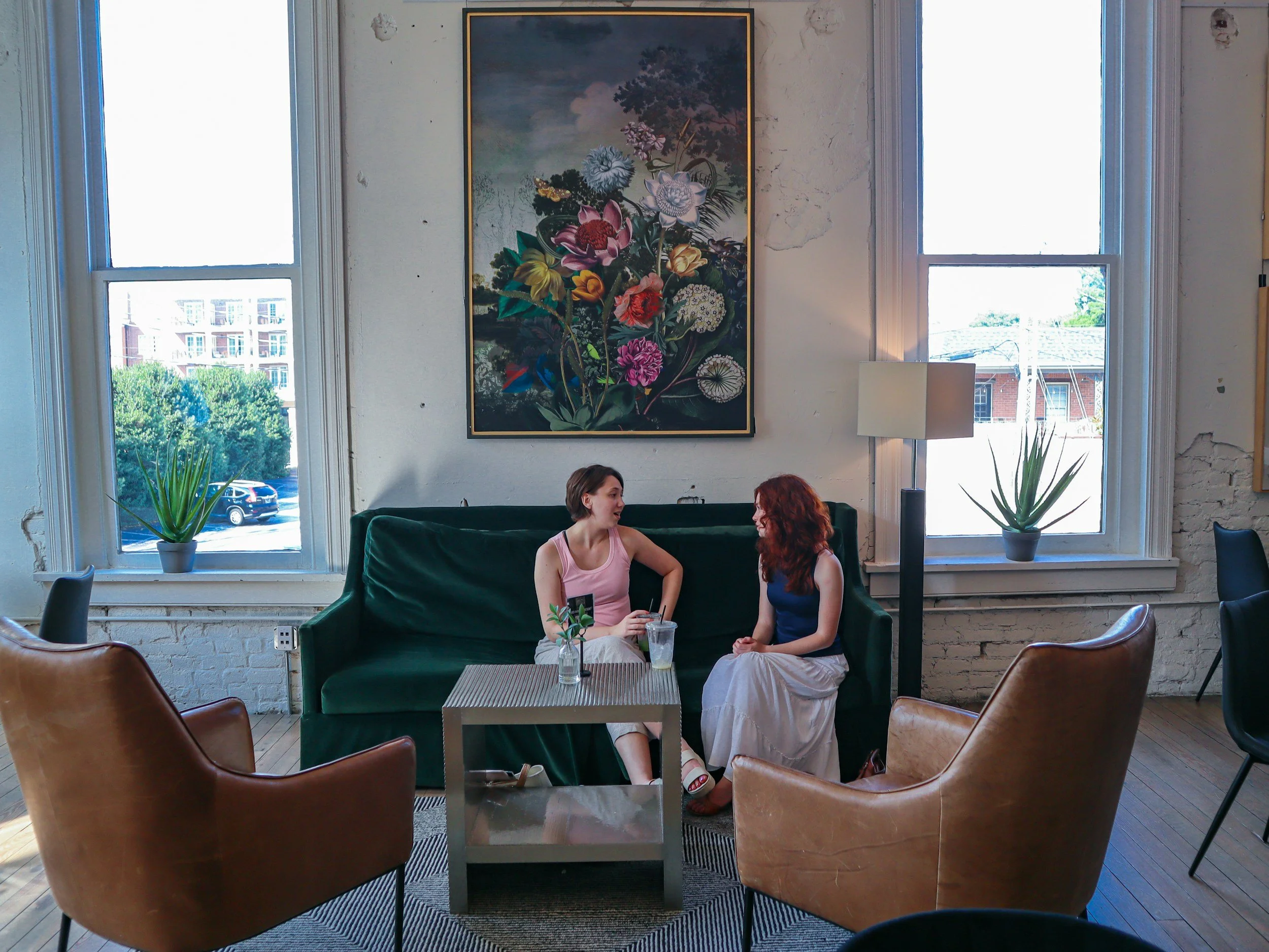 Two women sitting on a green velvet sofa having a conversation in a room with large windows, framed by white brick walls, with a large floral painting and potted plants nearby.