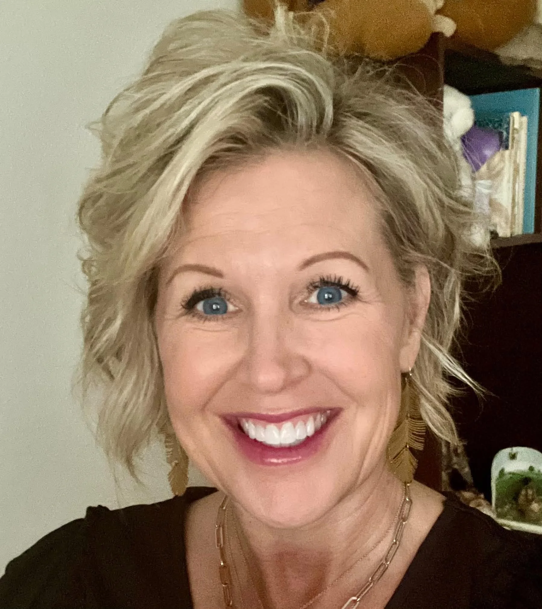 Close-up of a woman with blonde, curly hair and bright blue eyes, smiling showing her teeth, wearing a black top, layered gold necklaces, and earrings. In the background, there are bookshelves and a wall.